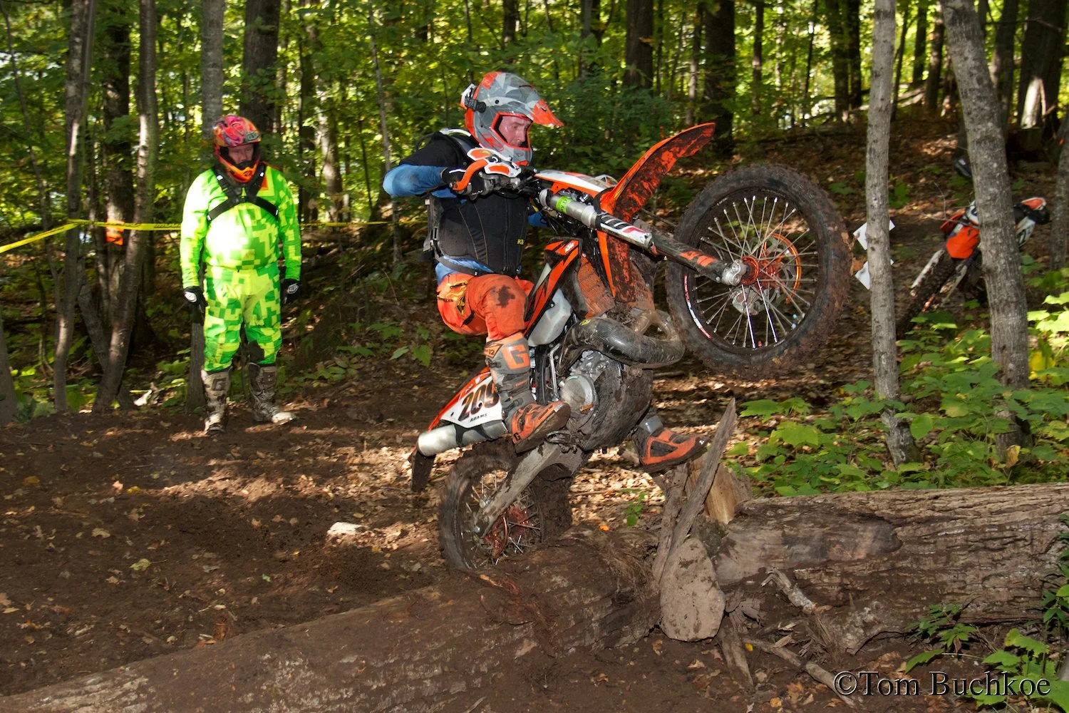 An off-road motorcycle rider wearing an orange helmet, blue jacket, and orange pants maneuvers over a fallen log on a dirt trail in a forest. A person in neon green gear observes nearby.