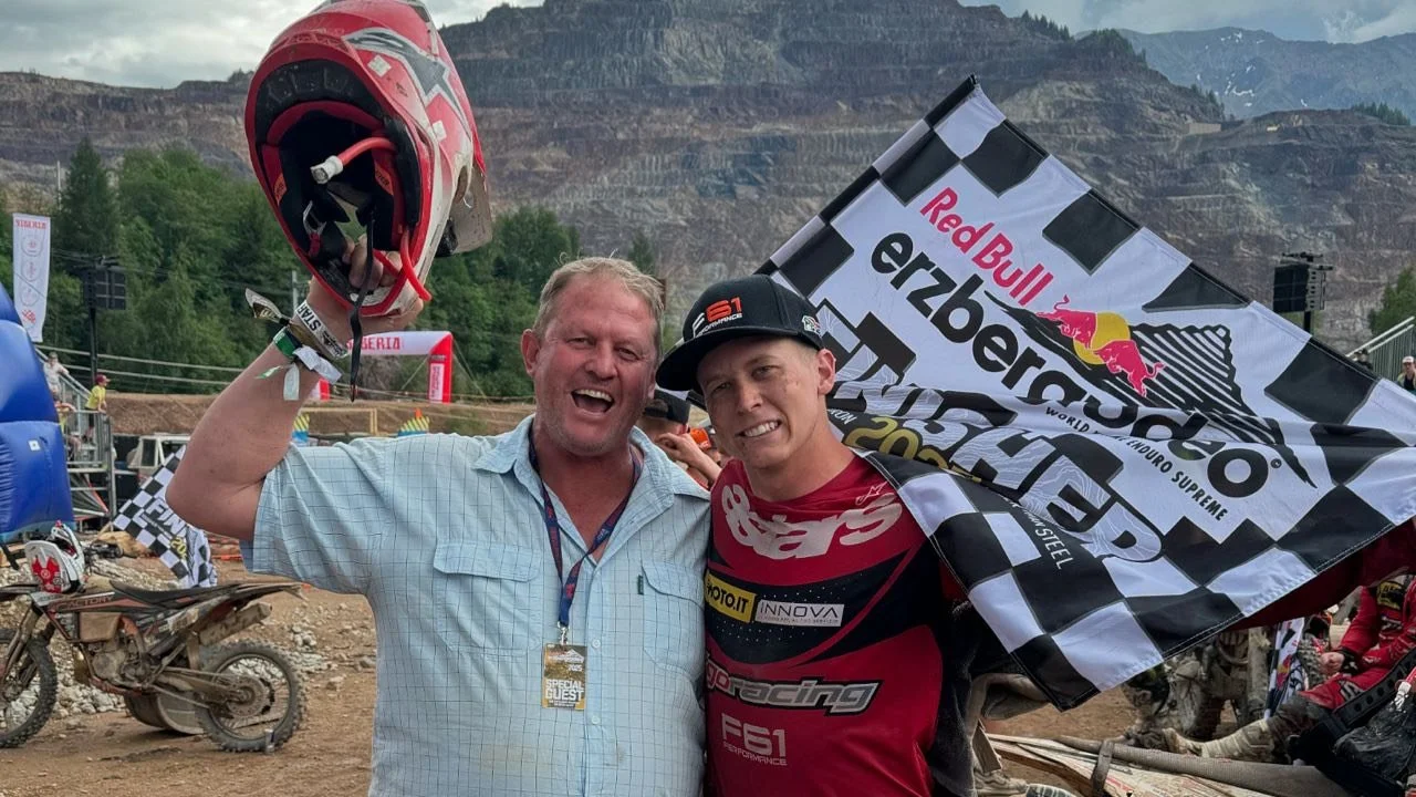 A man and a young motocross rider celebrating after a race, with the rider holding a flag that says 'Red Bull Erzbergrodeo', in a dirt motocross track area with bikes and a mountainous background.