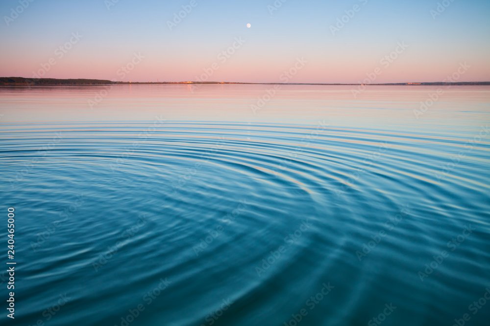 Calm body of water at sunset with visible ripples, pink and blue sky, and a small moon in the sky.