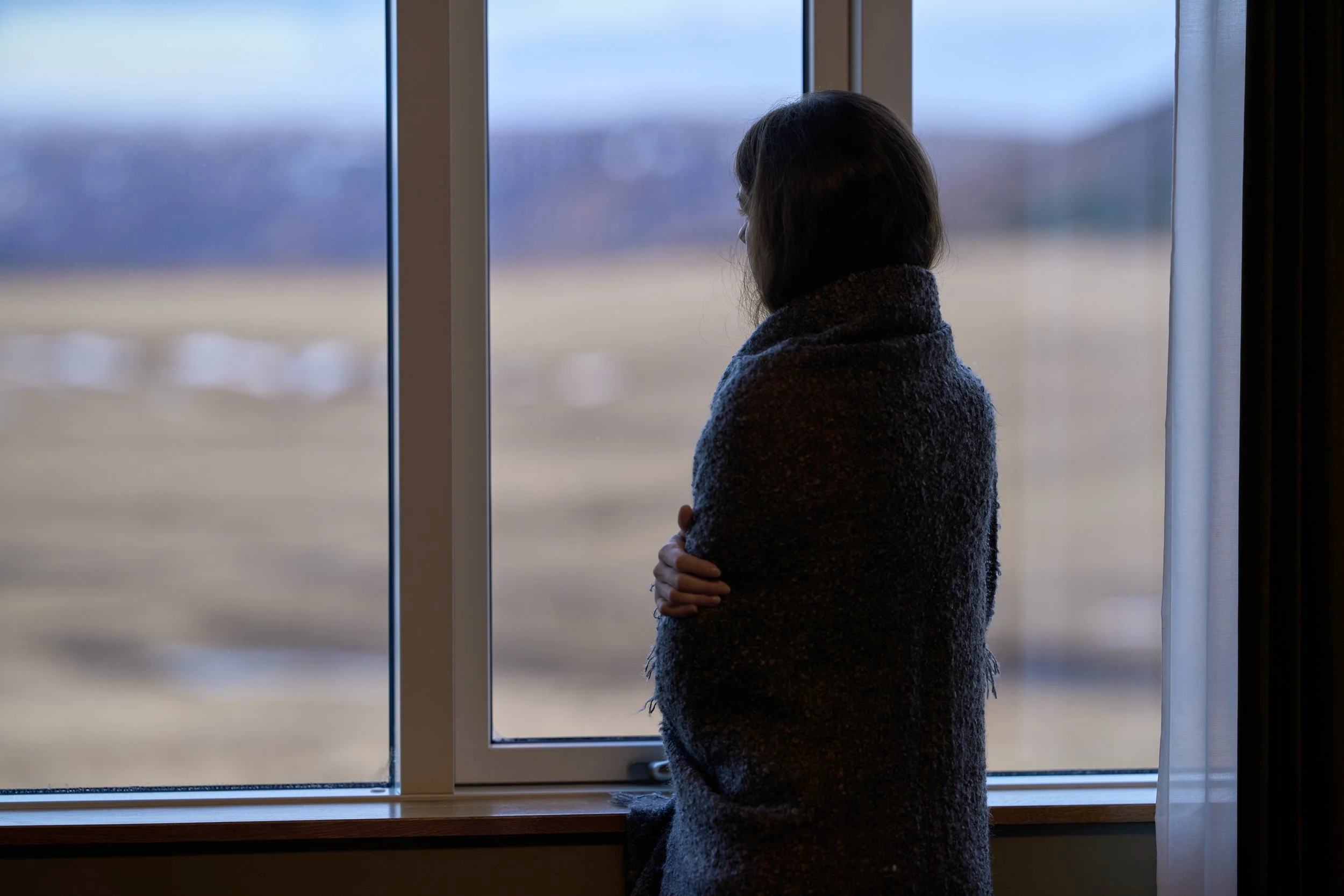 A woman standing near a window, looking outside with her arms crossed, during daytime.
