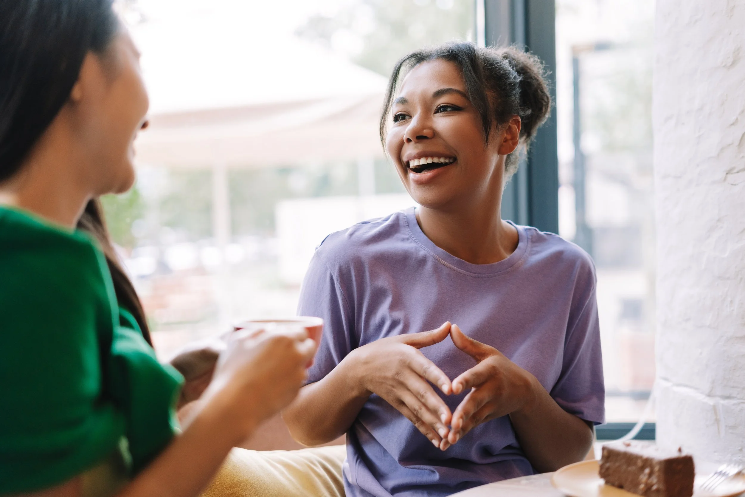 Two women are in a conversation, one smiling and the other partially visible, both sitting by a large window.