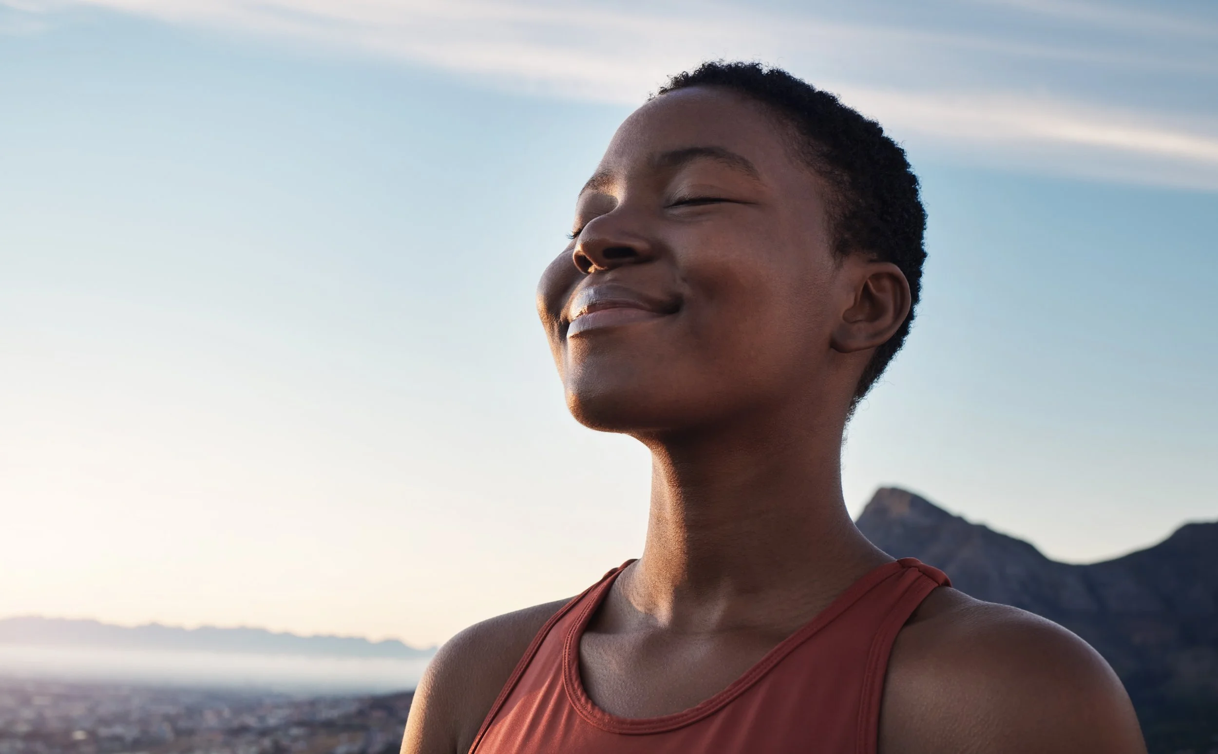 A person with short dark hair and medium skin tone is smiling with closed eyes, standing outdoors with a mountain in the background and a bright sky.