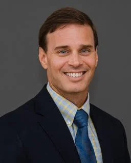 A smiling man in a dark suit, light blue shirt, and blue tie, standing against a gray background.