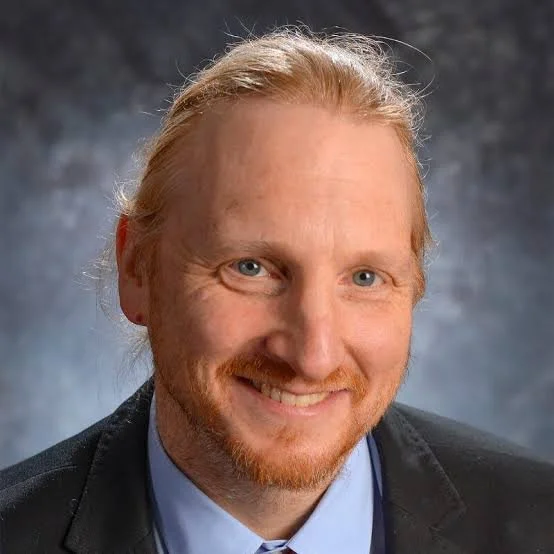 A man with red hair and beard, smiling, dressed in a dark blazer and light blue shirt against a dark background.