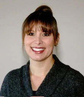 Portrait of a smiling woman with brown hair and bangs, wearing a dark gray top, against a plain background.