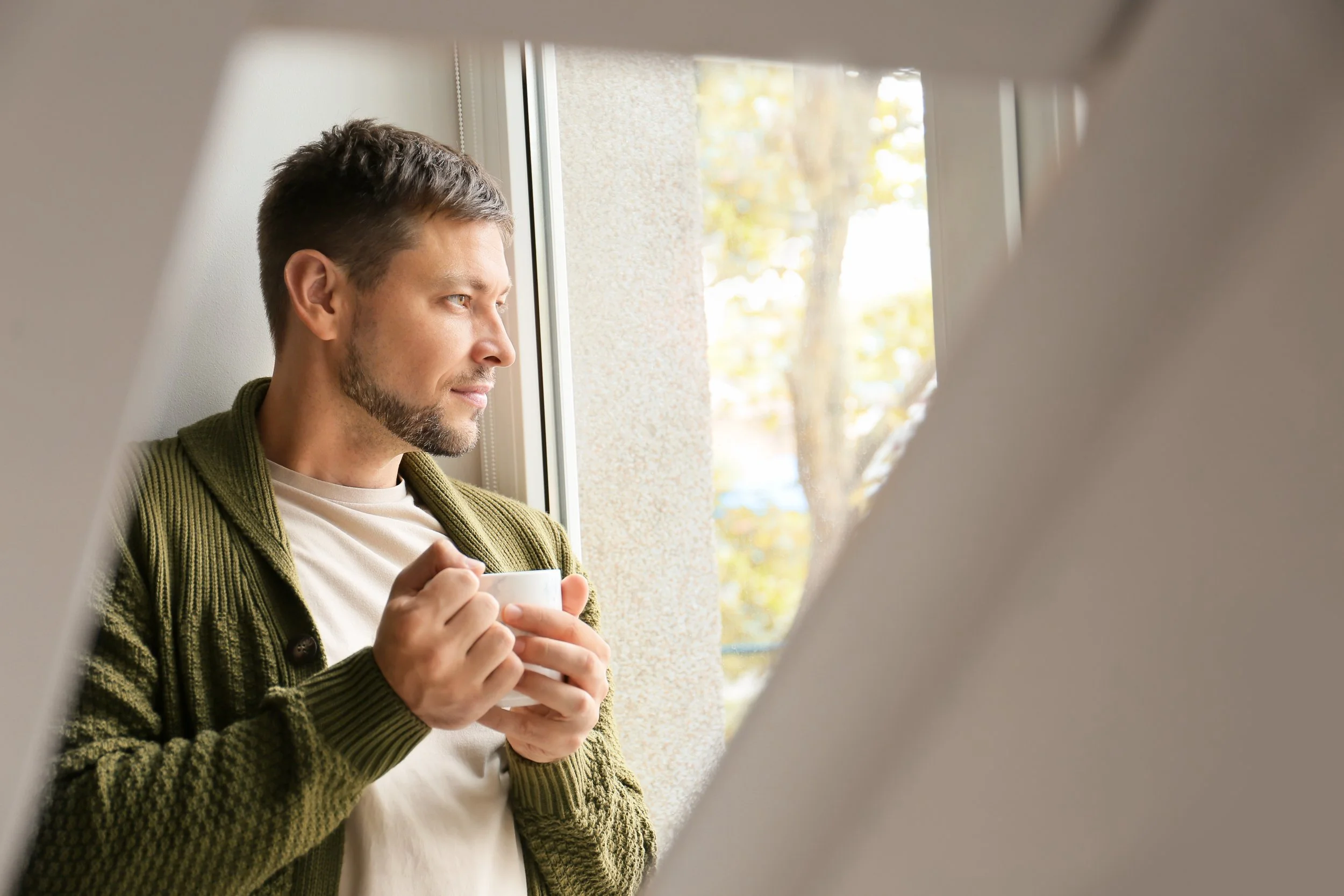 A man with short brown hair and a beard holding a white mug, standing next to a window with a view of trees outside.