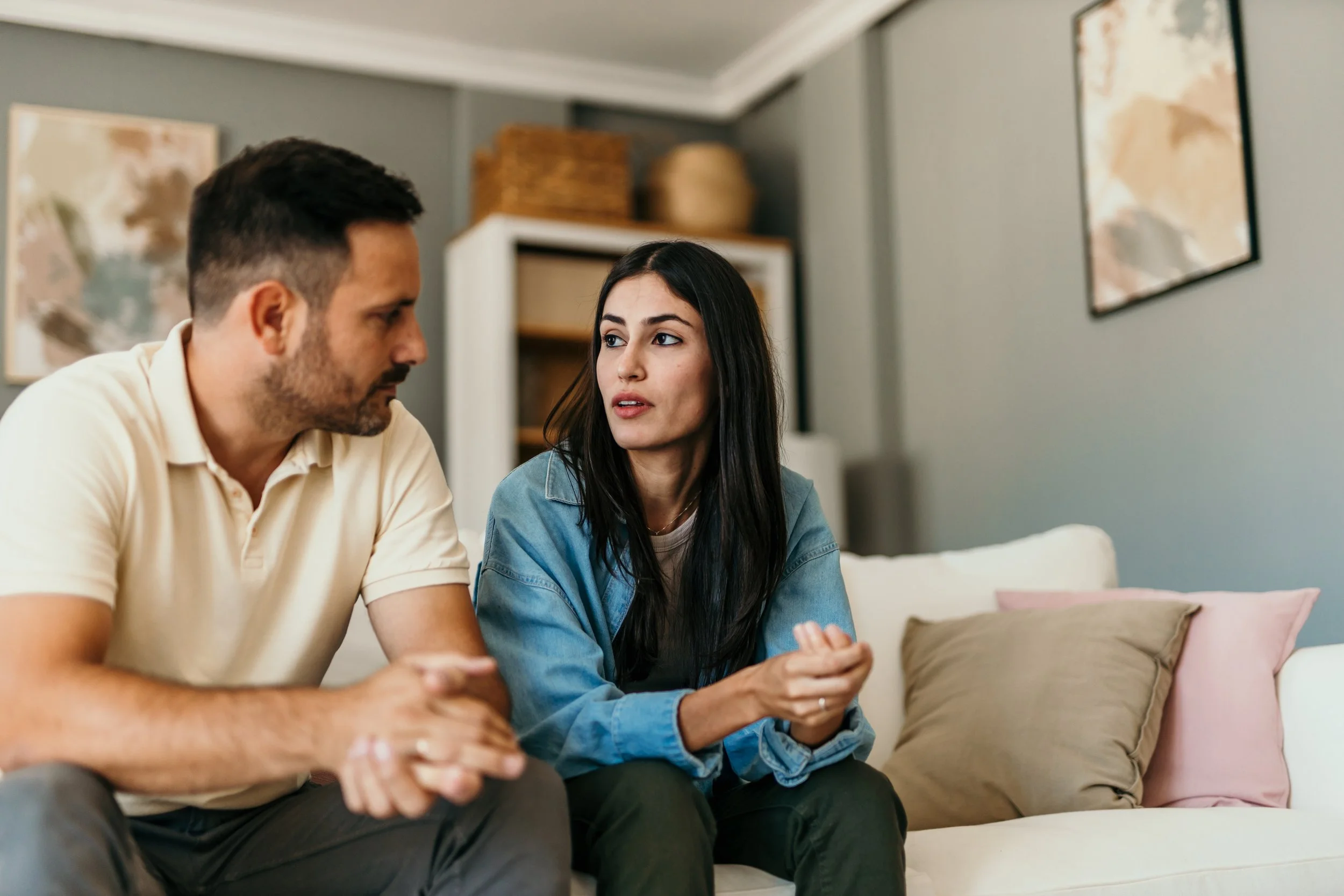A man and woman sitting on a couch engaged in a conversation indoors, with decorative artwork on the wall behind them.