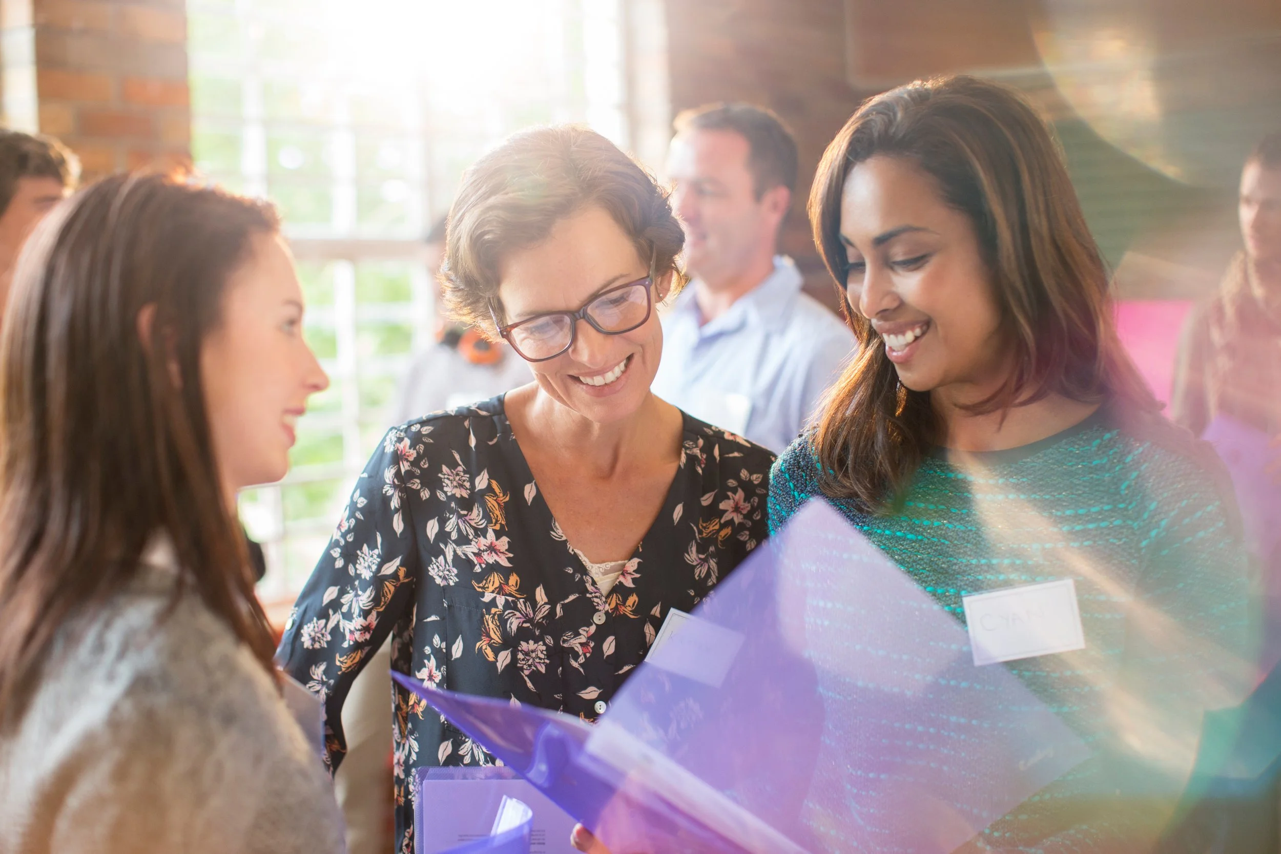 Group of people smiling and looking at a colorful display or project at a social or professional gathering in a bright room with large windows.