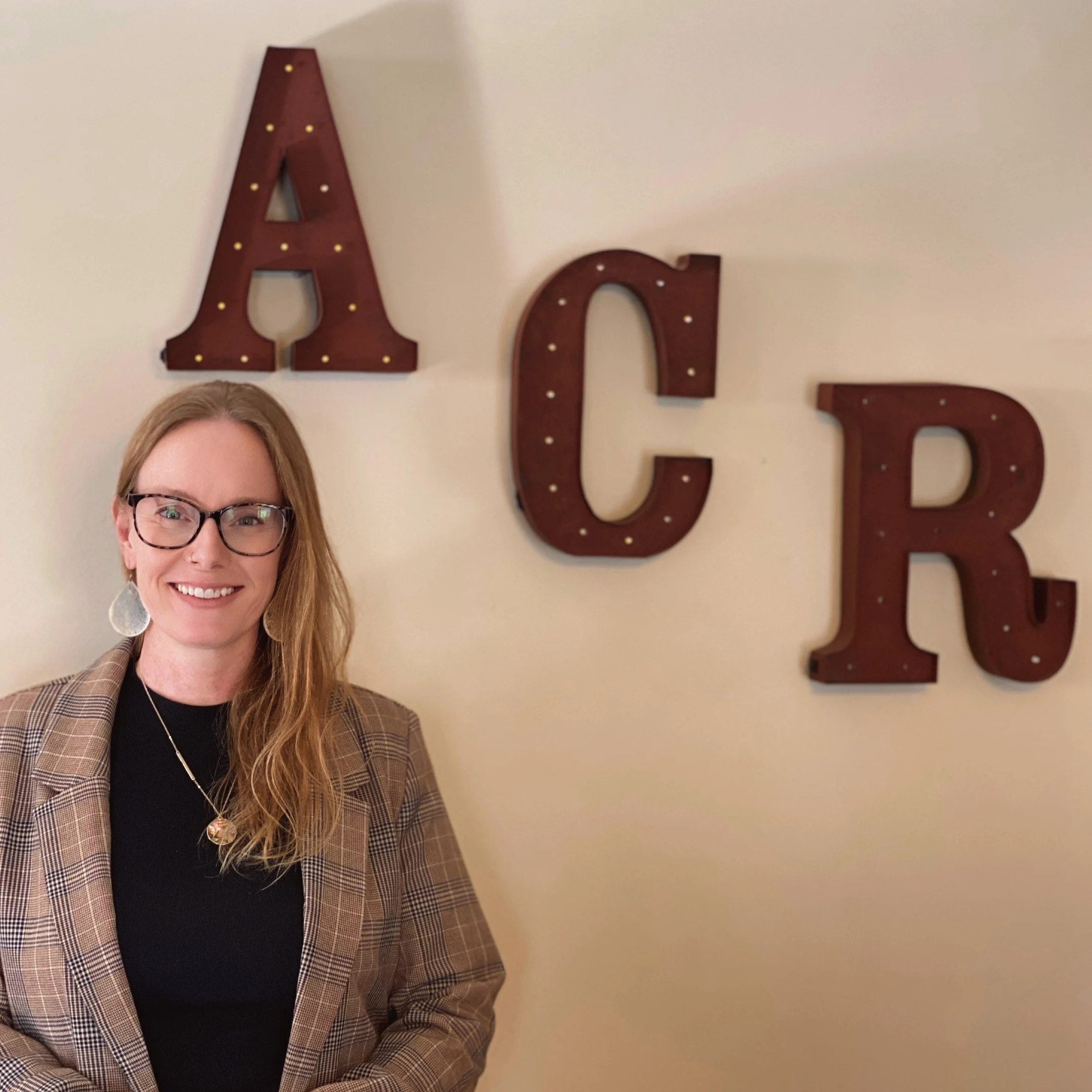 Partial view of a woman with glasses smiling in front of a wall with large decorative letters spelling 'ACE'.