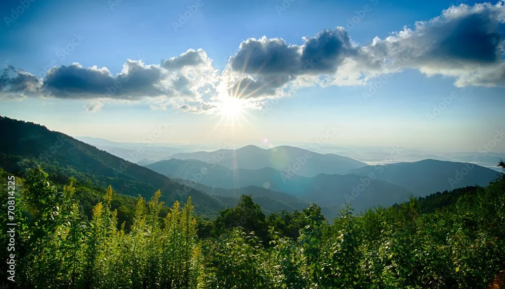 Sun shining through clouds over green mountains and hills with lush vegetation.