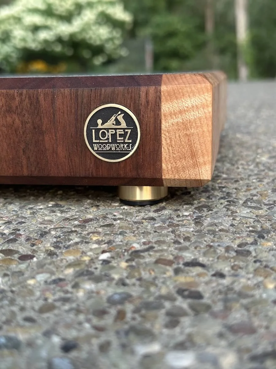 Close-up of a wooden object with a Lopez Woodworks metal badge, positioned on a gravel surface outdoors with blurred greenery in the background.