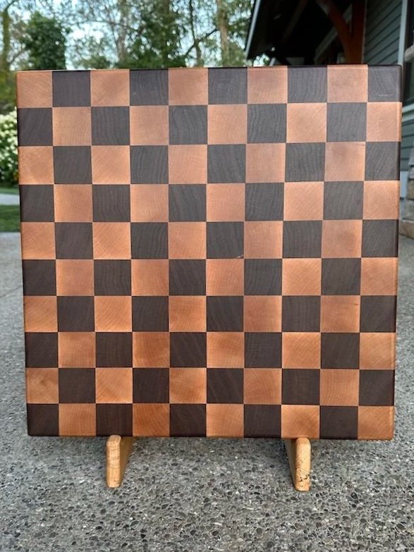 A wooden chessboard with alternating light and dark squares, standing on a small wooden easel outside on a driveway with trees and a house in the background.