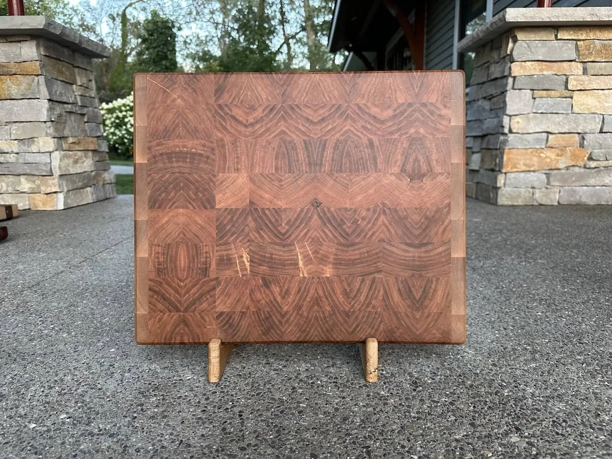 Square wooden cutting board with a decorative grain pattern, placed on a small wooden stand outdoors on a concrete surface with stone columns and a house in the background.