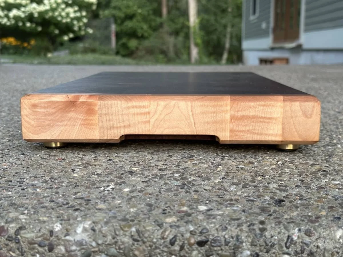 A wooden electronic scale on a gravel surface outdoors, with a house and trees in the background.