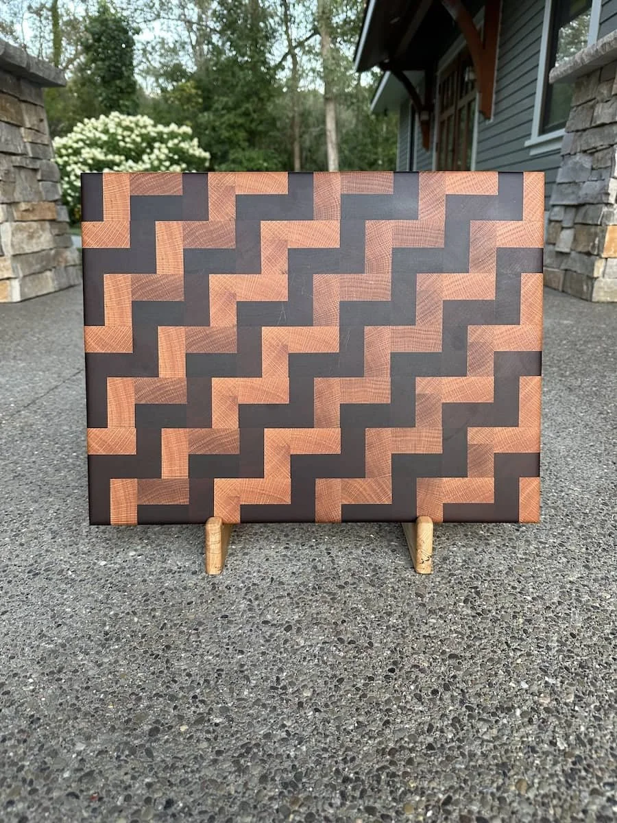 A geometric patterned wooden box with a zigzag design, standing on small wooden legs on a concrete surface outside a house with a stone wall and trees in the background.