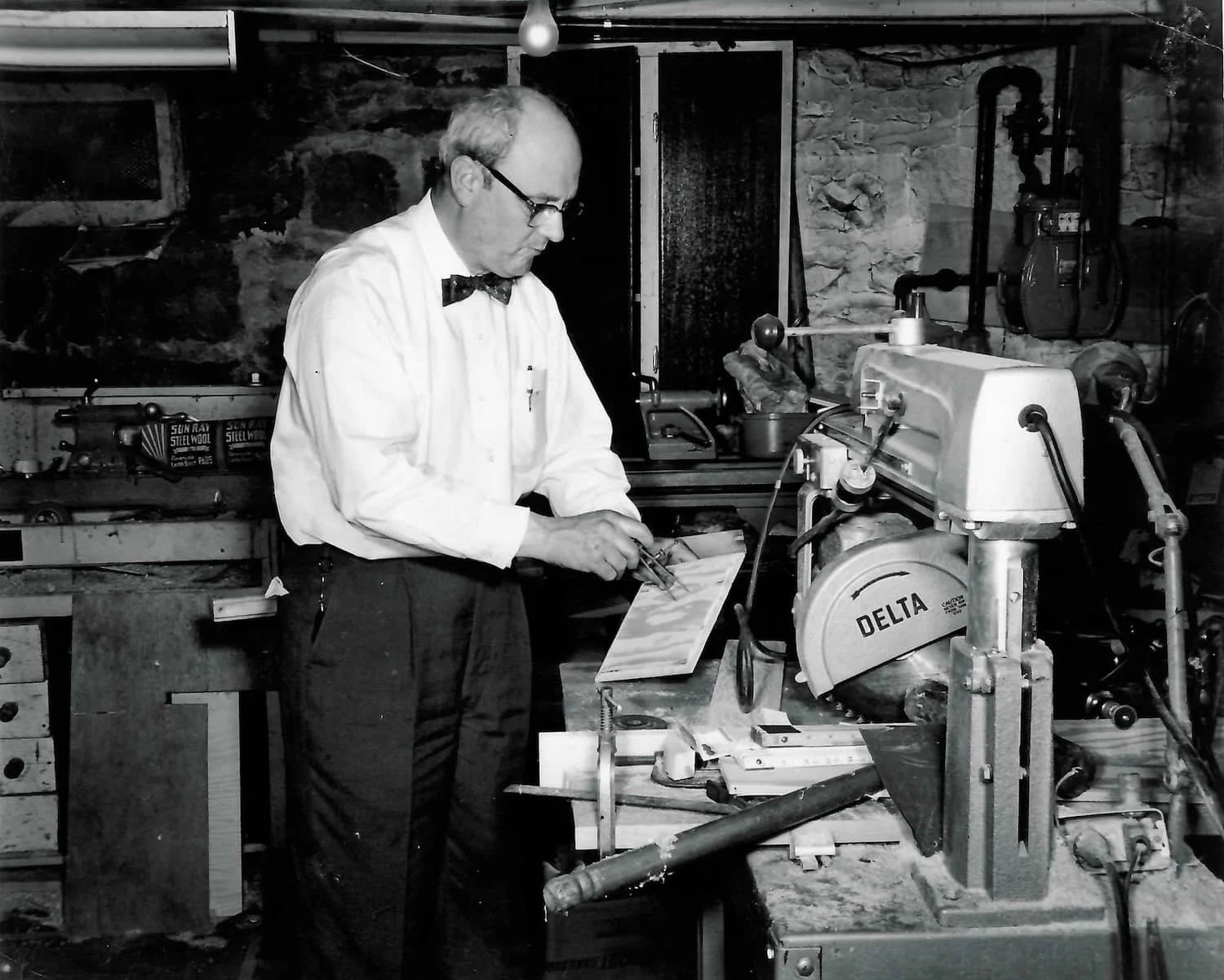 Historical photo of our grandfather in a white shirt, bow tie, and glasses working with a piece of equipment in a workshop with stone walls.