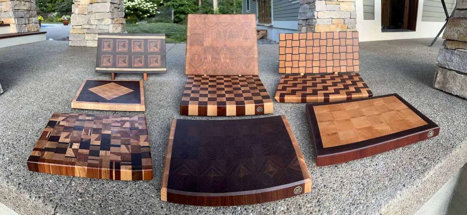 Collection of eight wooden cutting boards with various geometric patterns, arranged on a concrete outdoor surface near a house with stone pillars and a door.