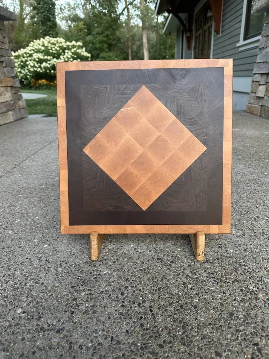 A wooden chessboard with a dark background and light wood squares, placed outdoors on a small wooden easel. In the background, there is landscaping with green trees and a house with gray siding.