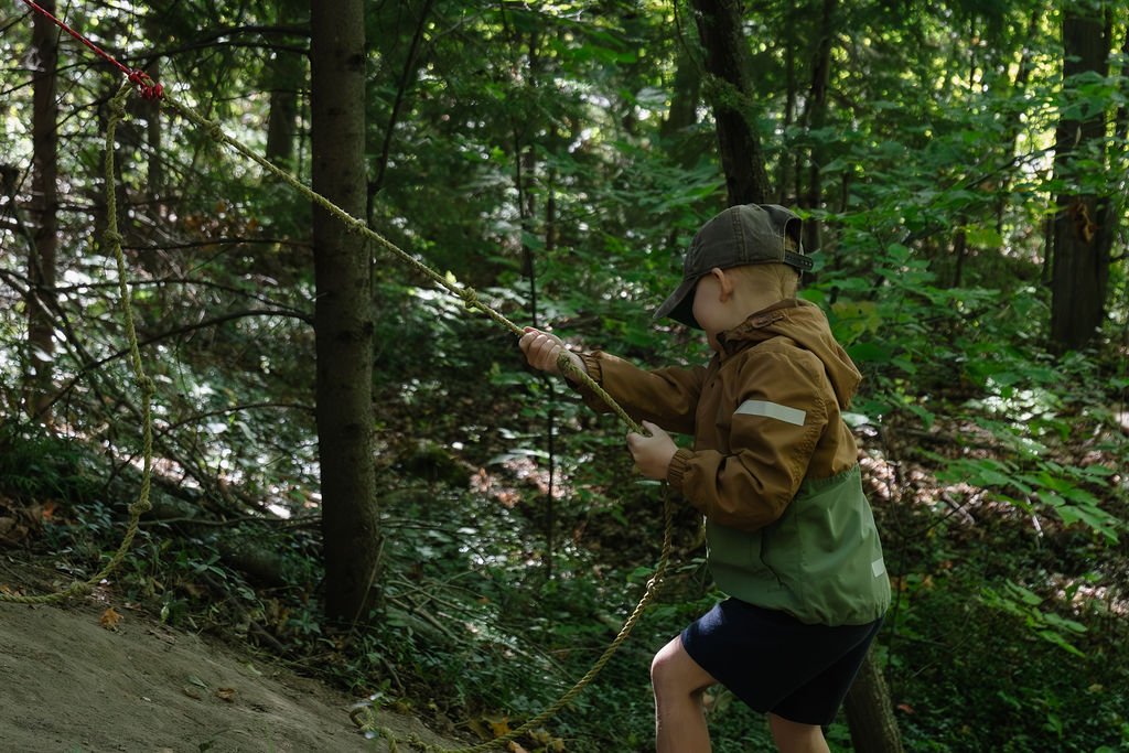 A young boy wearing a brown jacket, green pants, and a green cap skates on a slackline in a wooded area.