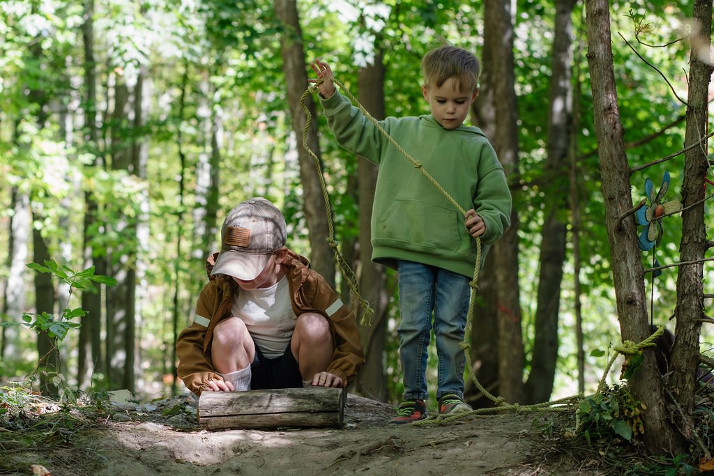 Two young boys exploring in a wooded forest, one sitting with his head bowed and the other holding a rope.