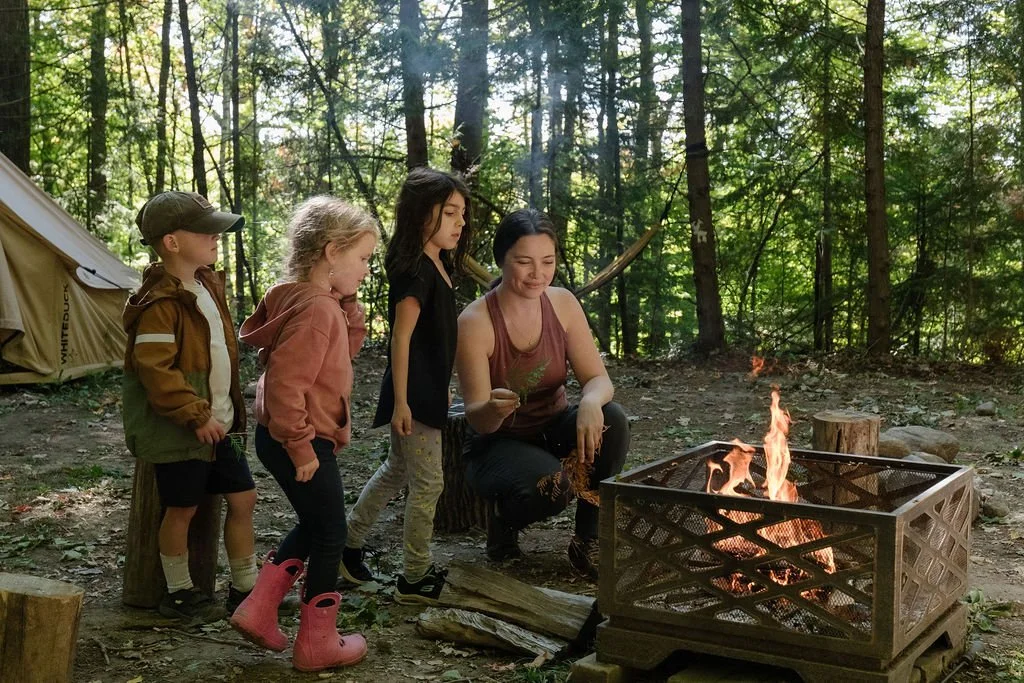 A group of four children and a woman in a forest clearing around a campfire. The children are standing, watching the fire, with a tent visible in the background.