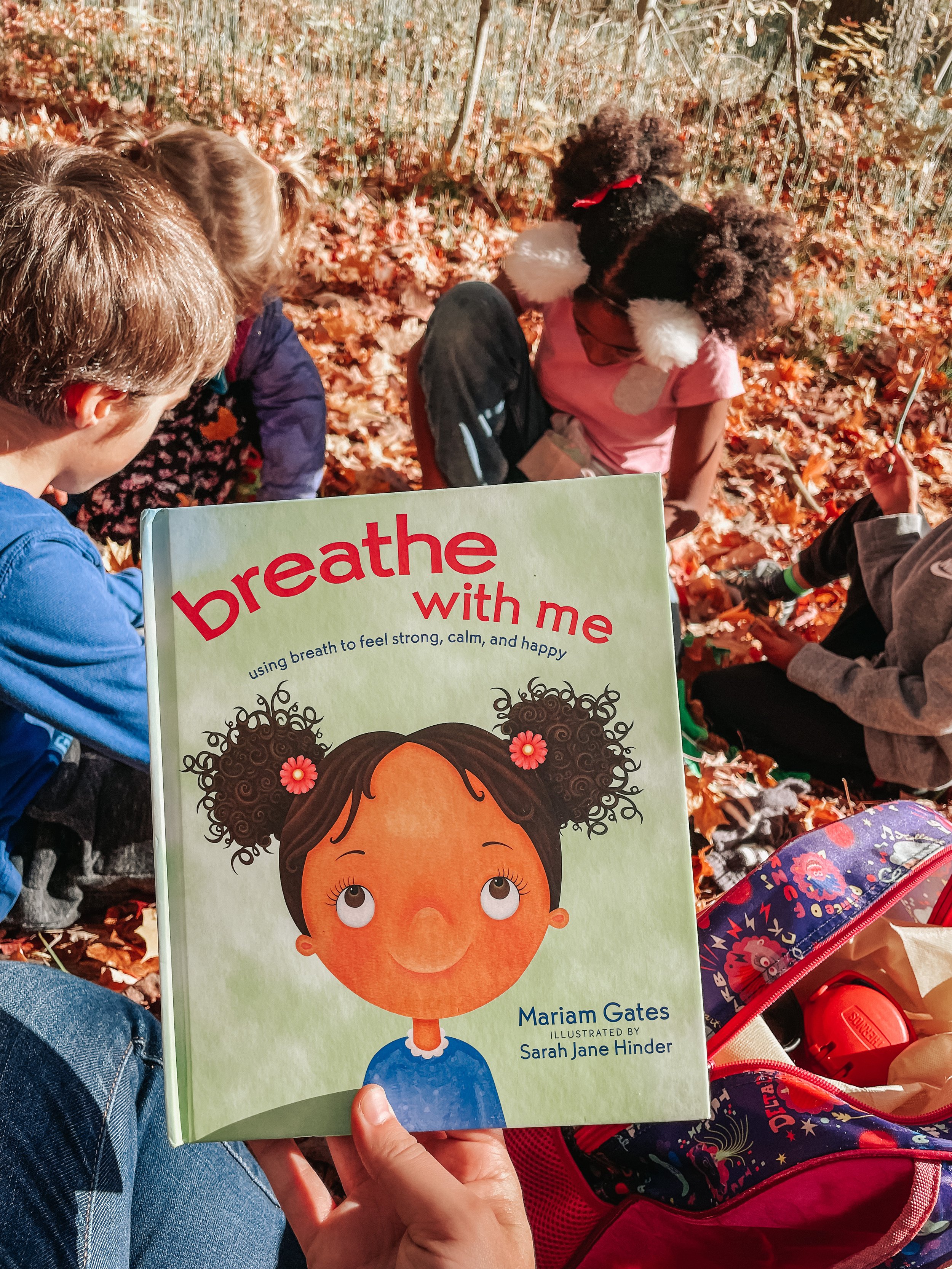 A person holding a children's book titled 'Breathe with Me' in front of a group of kids sitting on the ground in a fall forest with orange leaves. The book cover features a girl with curly hair and a smiling face.