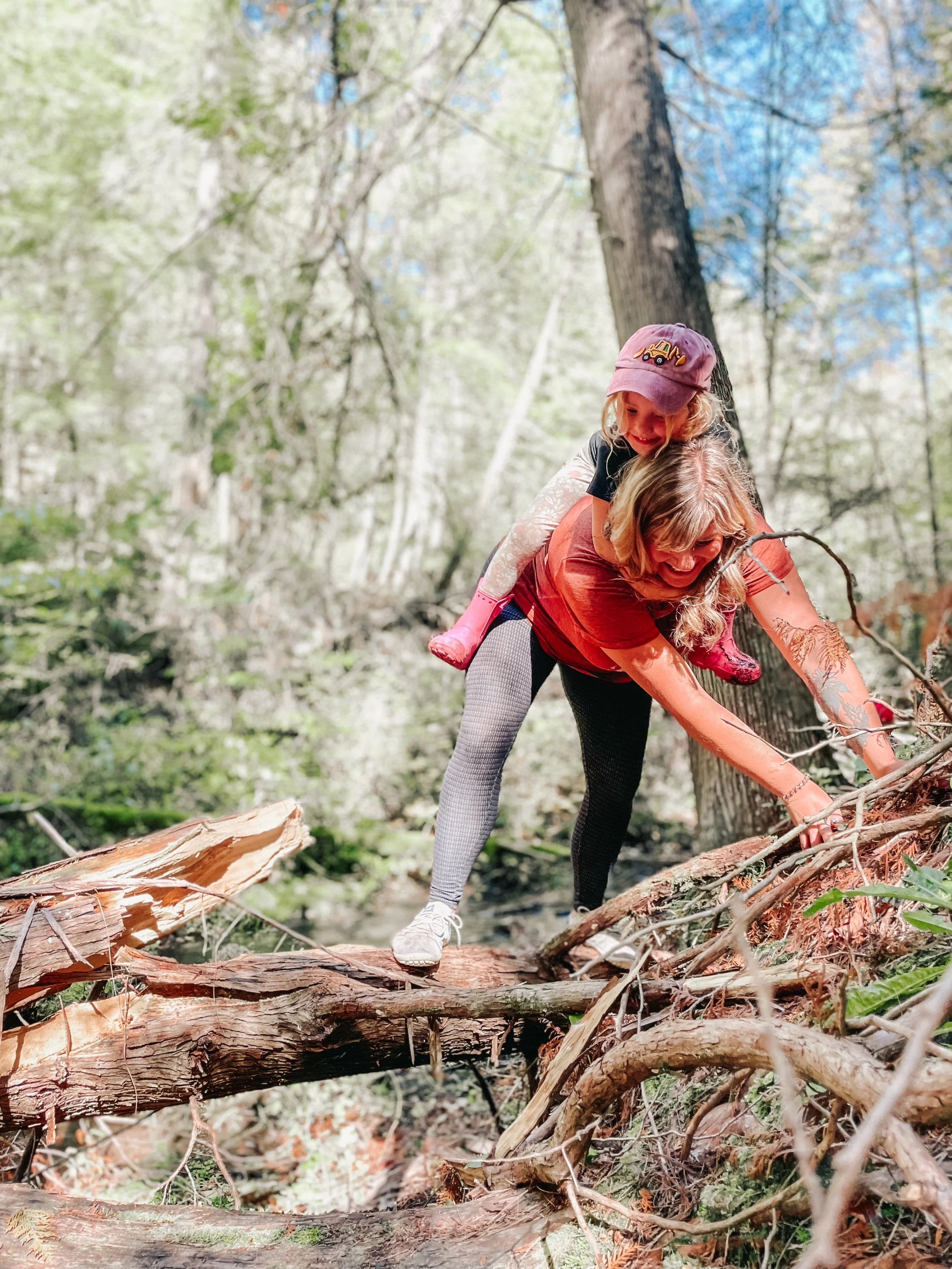 A woman and a young girl are outdoors in a forest, balancing on a fallen tree trunk. The woman is bending forward with a smile, supporting the girl on her back who is wearing a pink hat and pink boots.