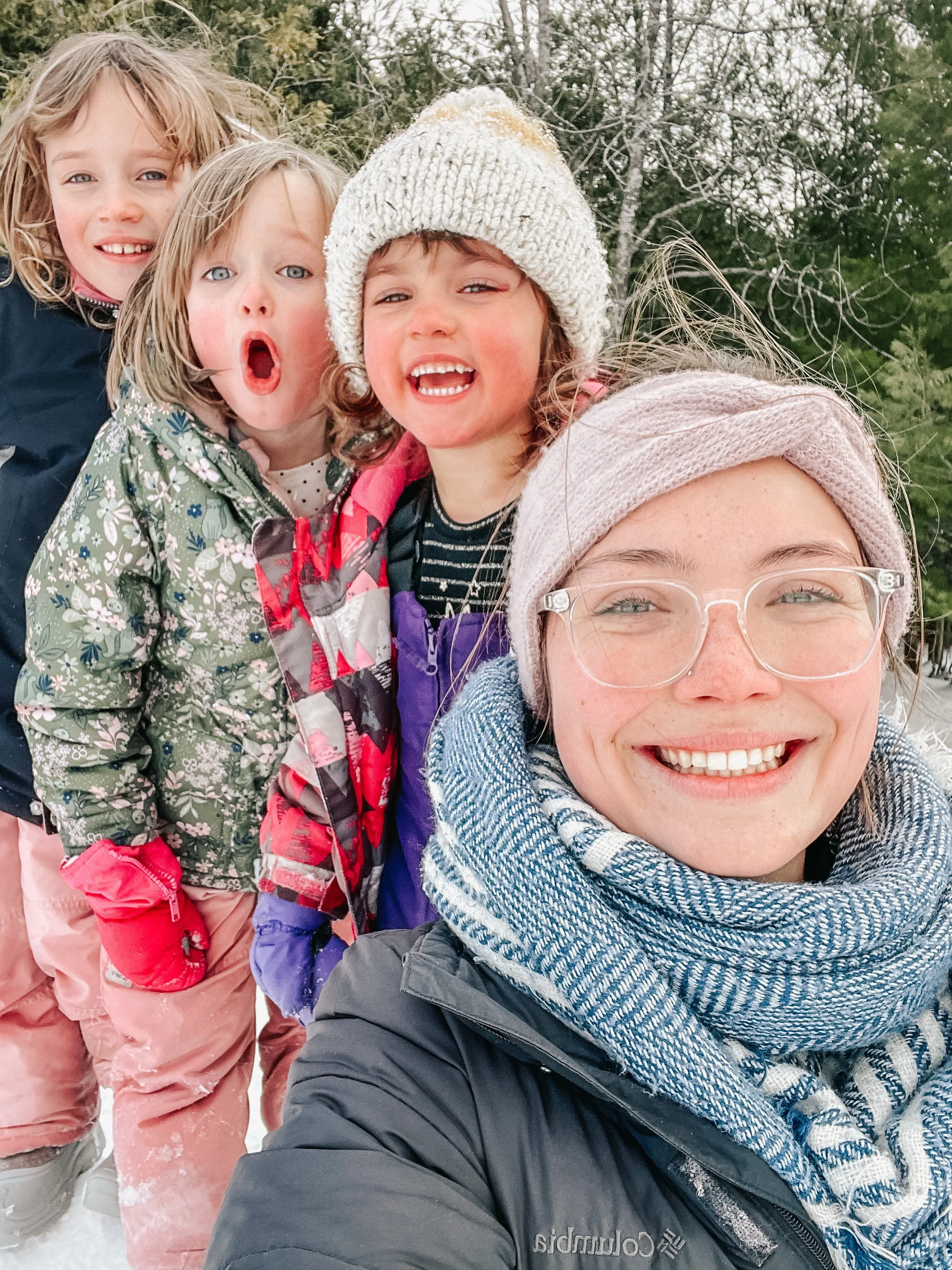 A smiling woman wearing glasses and a headscarf taking a selfie with four young girls dressed in winter clothing outdoors.