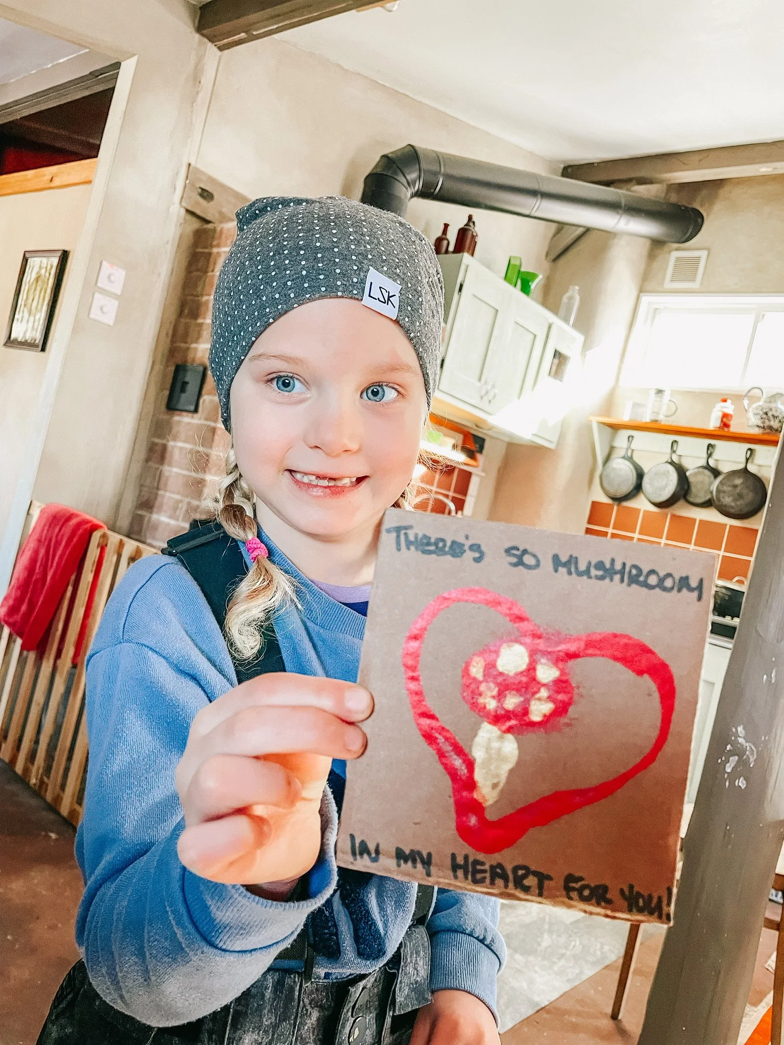 A young girl with blue eyes and blonde hair in a braid, wearing a gray beanie hat with a white label, holding a handmade card that says "There's so mushroom in my heart for you!" The card has a drawing of a mushroom inside a red heart.