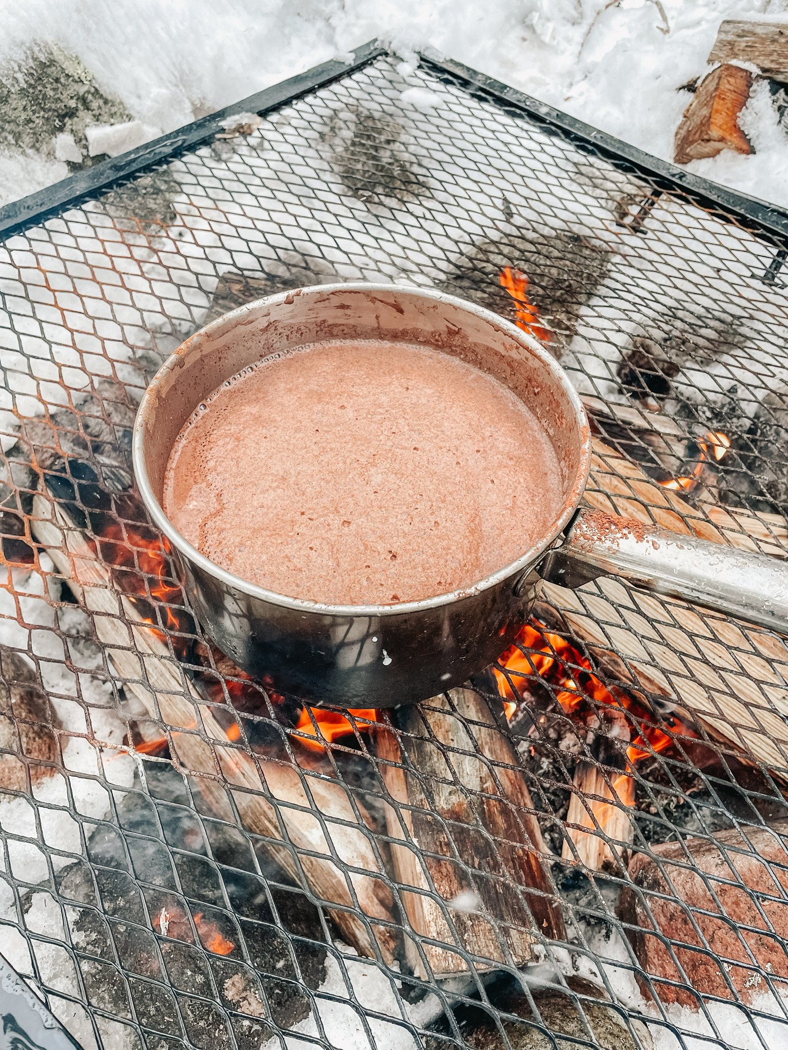 A pot of hot chocolate boiling over an outdoor campfire grate, with snow in the background.