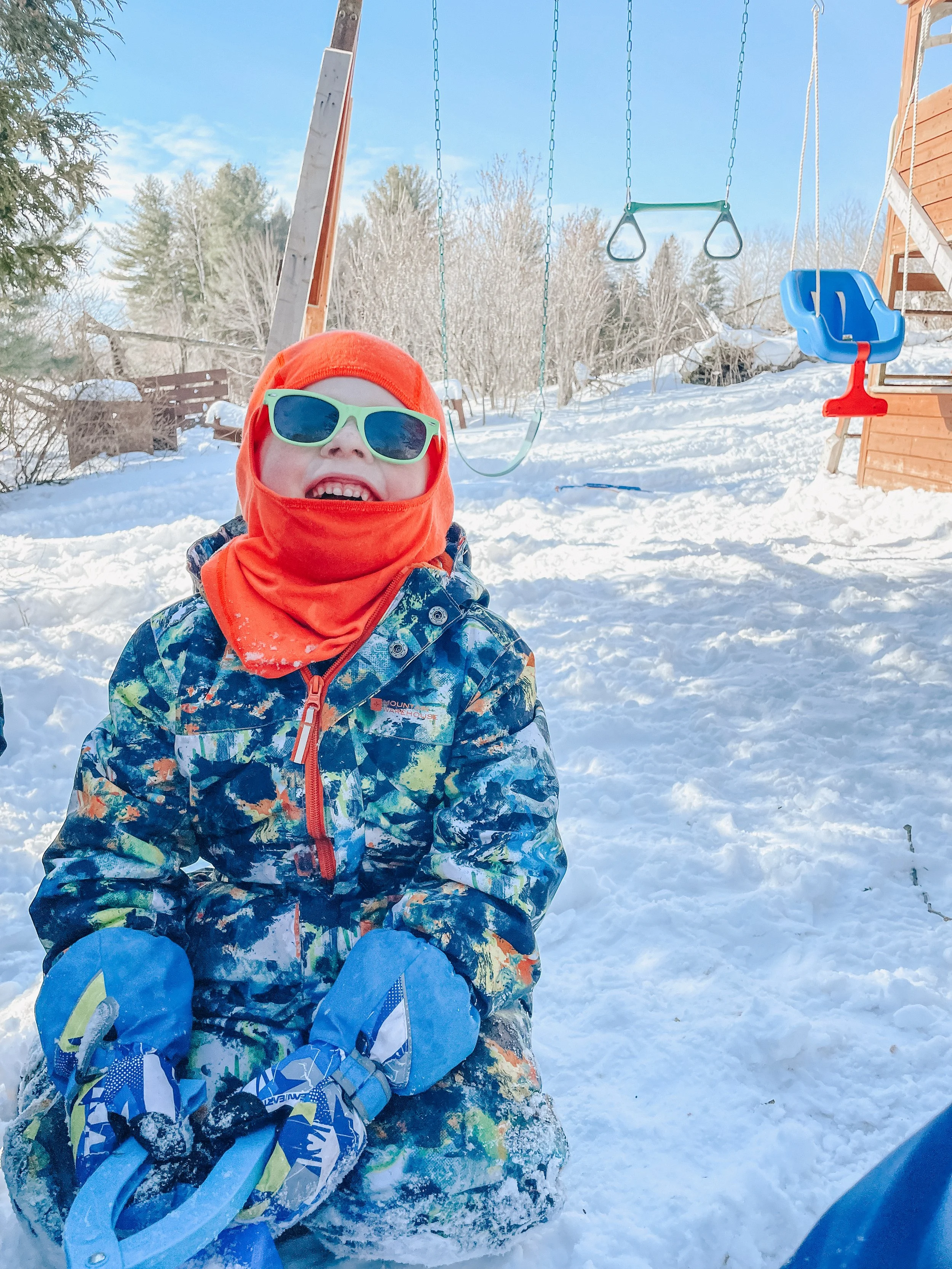 A young child in colorful winter gear, including sunglasses, a red hat and scarf, and a patterned snowsuit, playing in the snow outdoors on a sunny day. In the background, there is a swing set and a wooden slide.