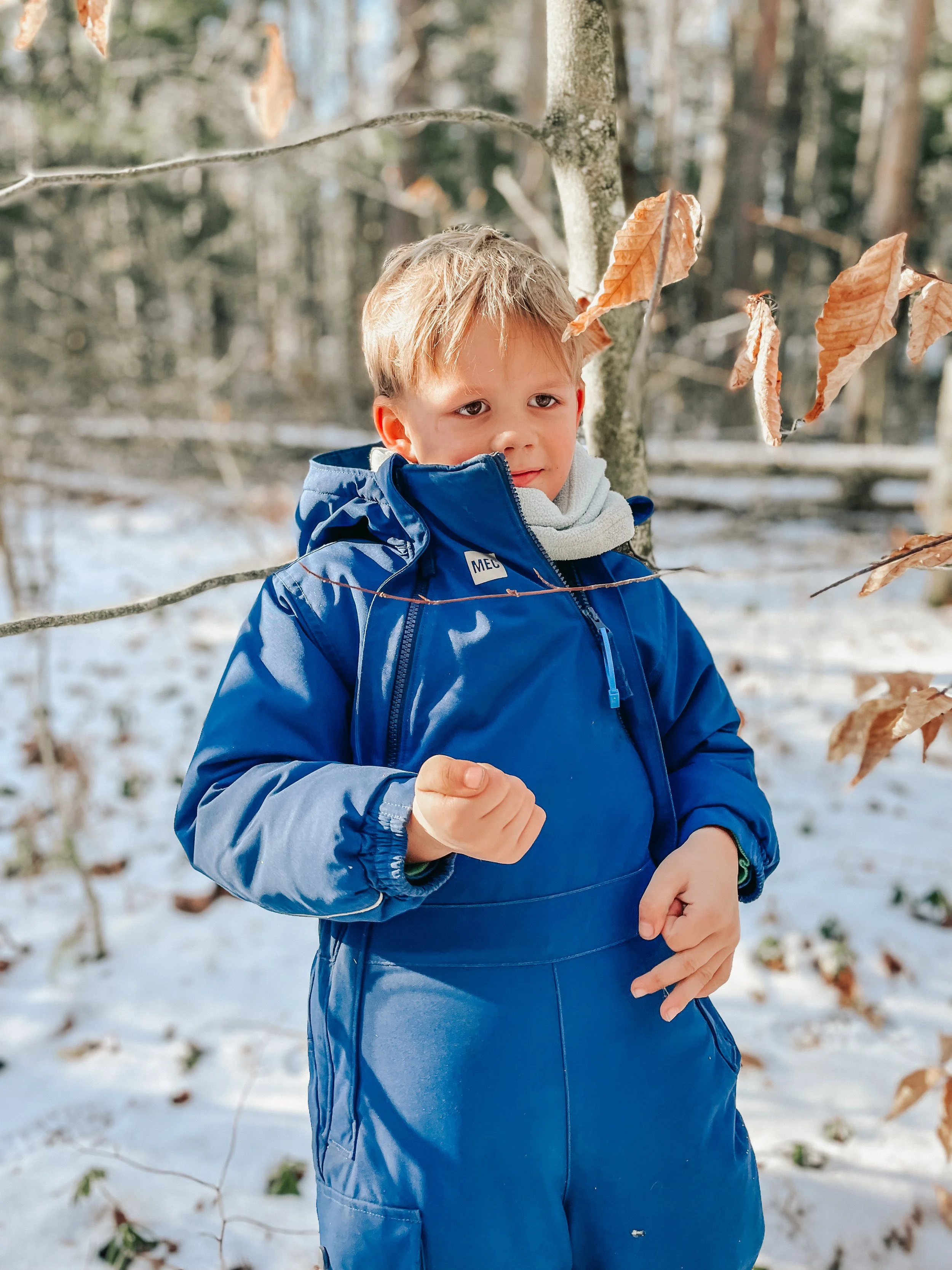 A young boy wearing a blue winter jacket and gray scarf standing outdoors in a snowy forest, looking concerned or thoughtful.