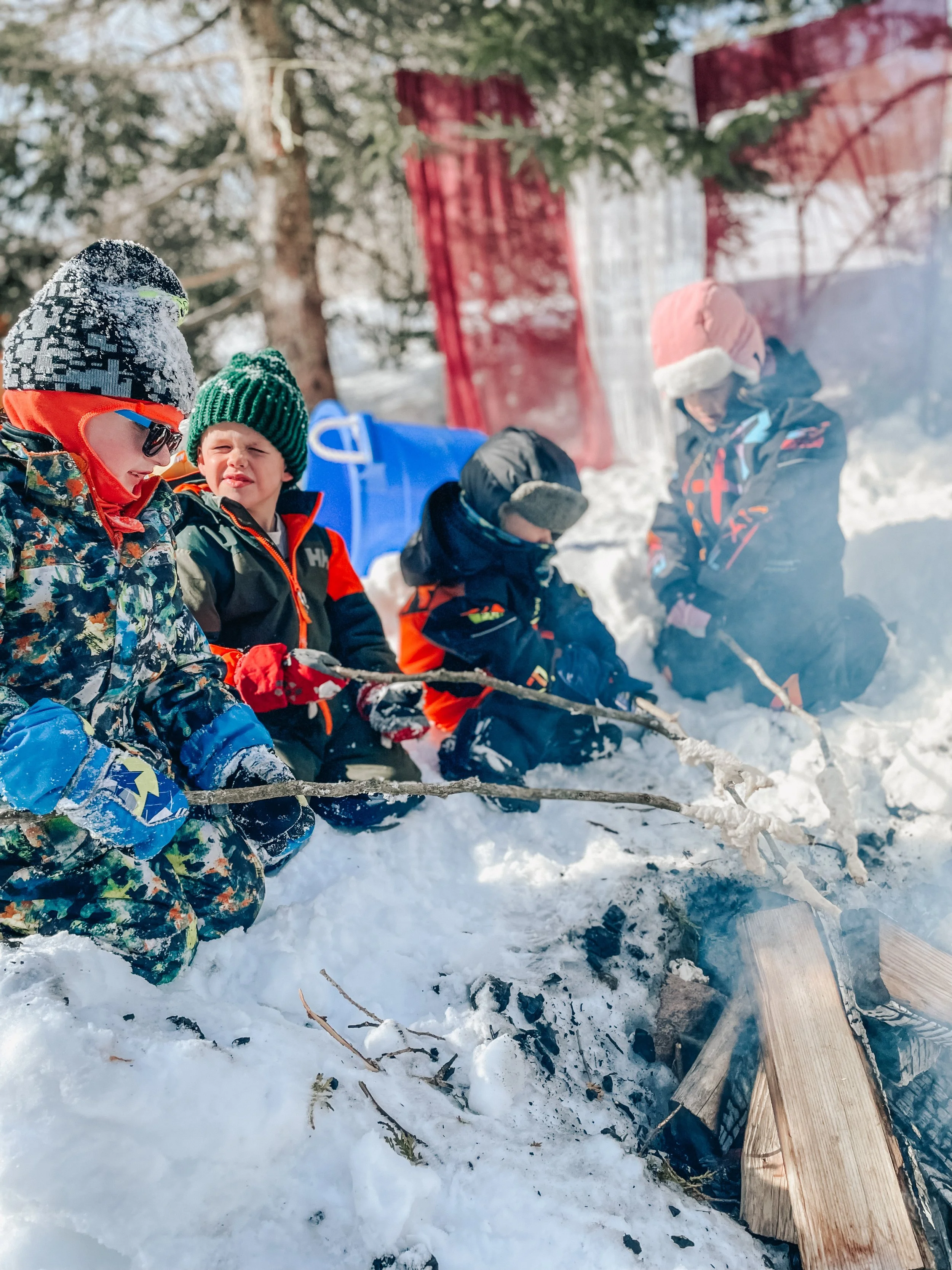 Four children sitting by a campfire in snowy outdoors, dressed in winter gear, with a red fence and trees in the background.