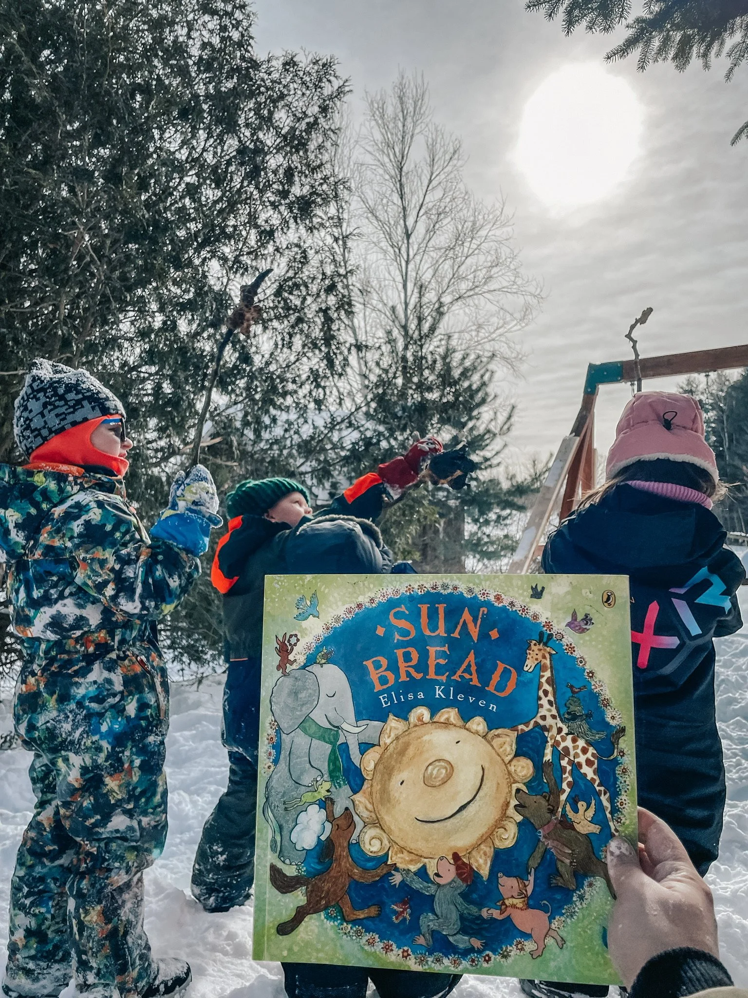A person holding a children's book titled 'Sun Bread' by Elisa Kleven in front of a group of children in winter clothing outdoors in a snowy landscape, with trees and a bright sun in the sky.