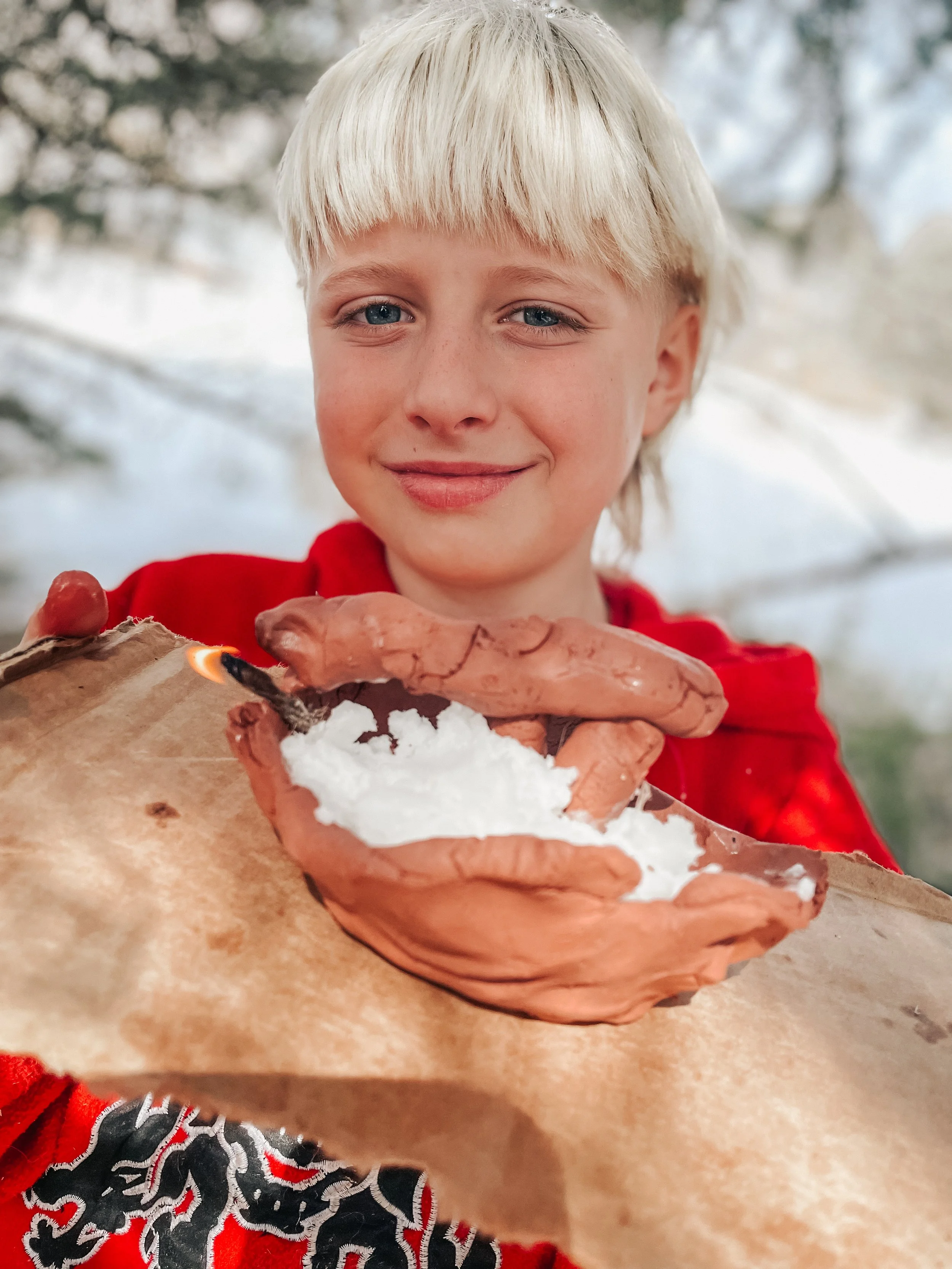 A young boy with blond hair and blue eyes holding a lit matchstick near a small pile of snow on a piece of brown paper, outdoors during winter.
