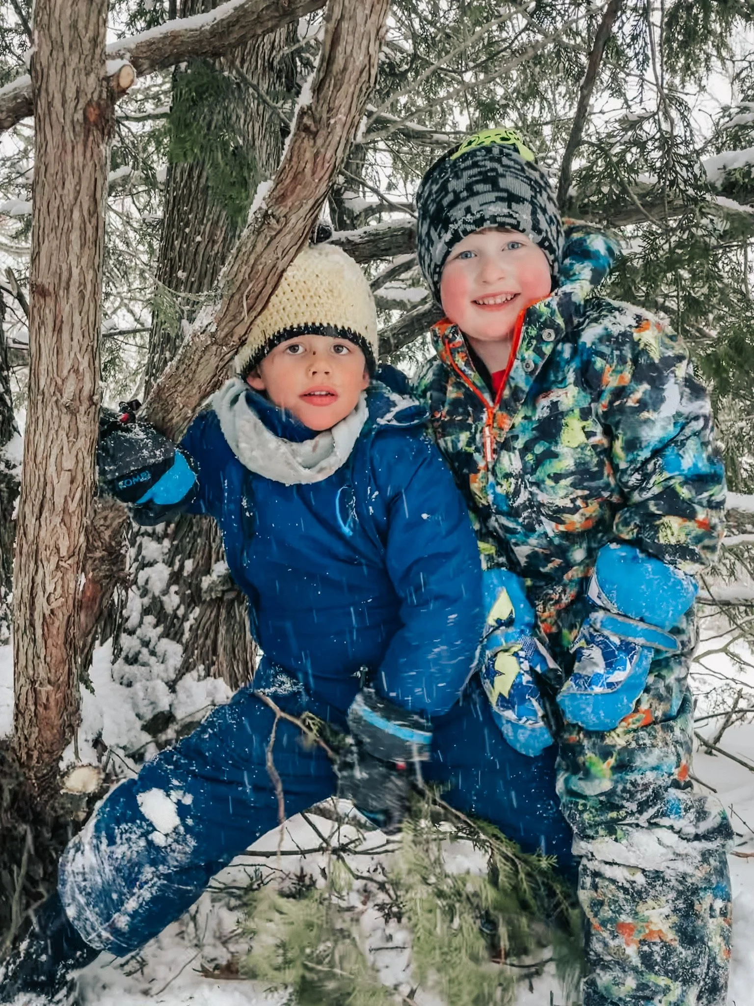 Two young boys in winter clothes playing in the snow near a tree, smiling and looking at the camera.