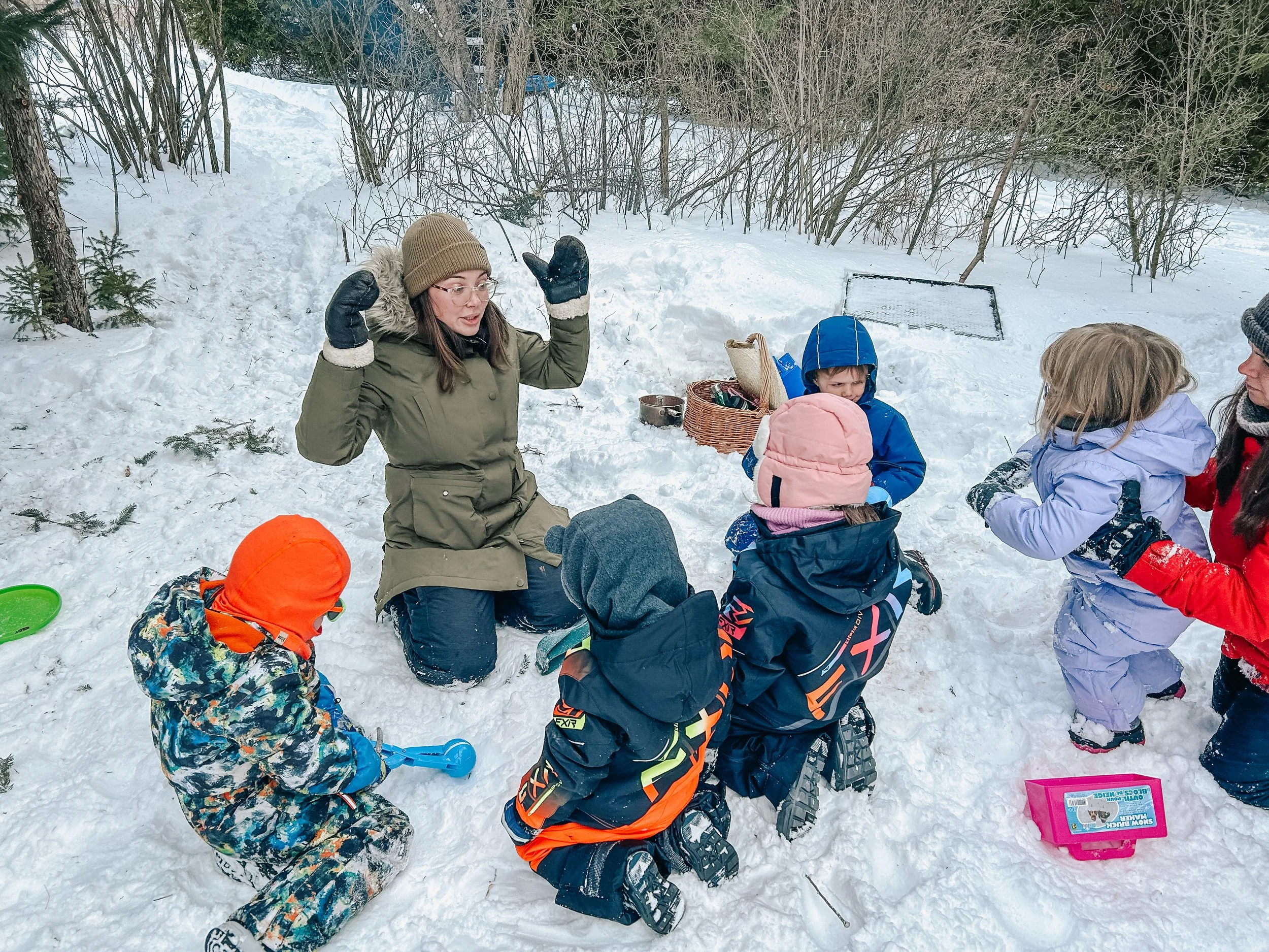 A woman in winter clothing teaches a group of children in snow, sitting and kneeling, surrounded by trees and snow-covered ground.