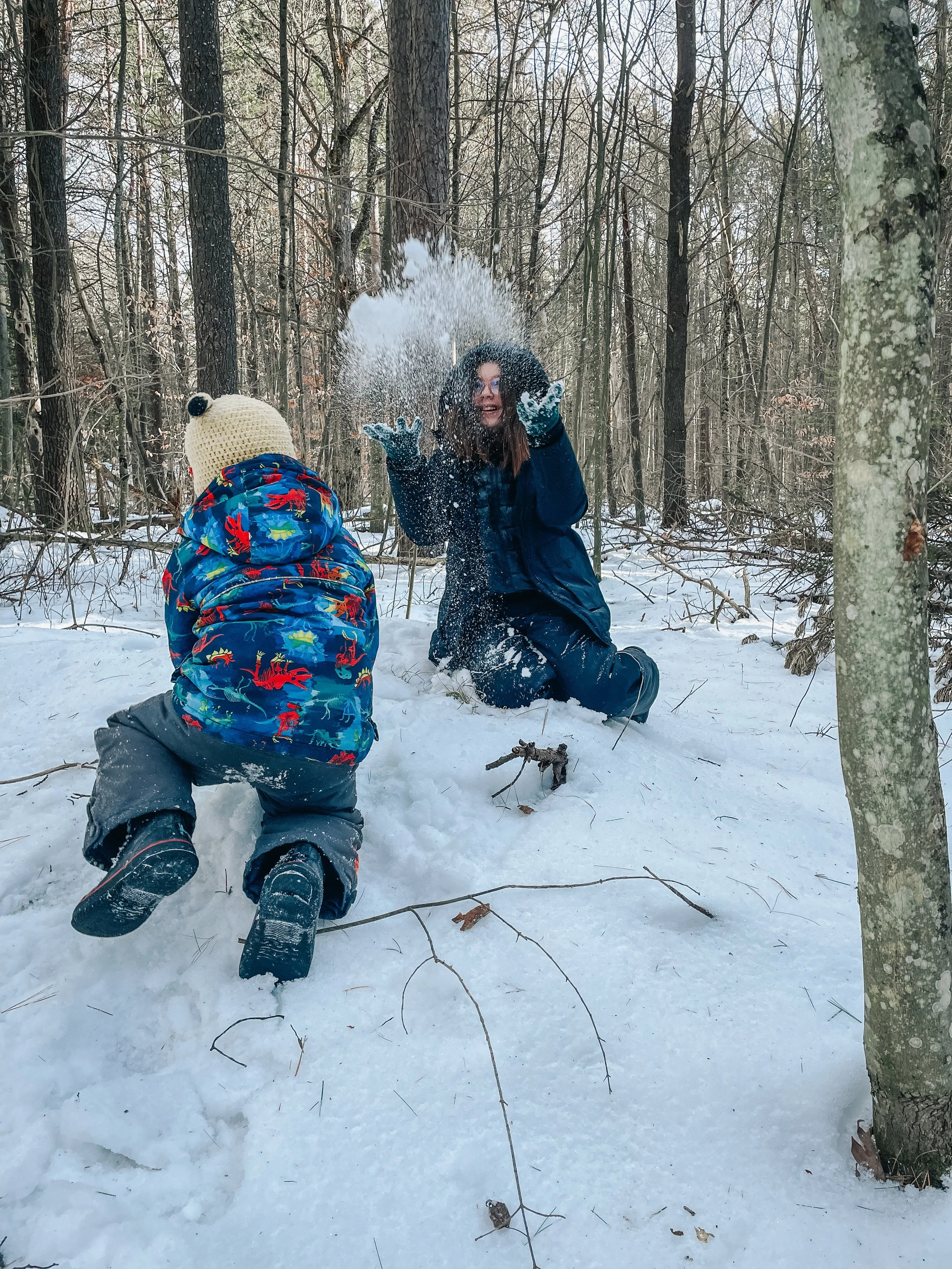 A woman and a young child playing in the snow in a forest, with the woman tossing snow into the air and the child kneeling in front, wearing a colorful jacket and a cream knit hat with a pom-pom.