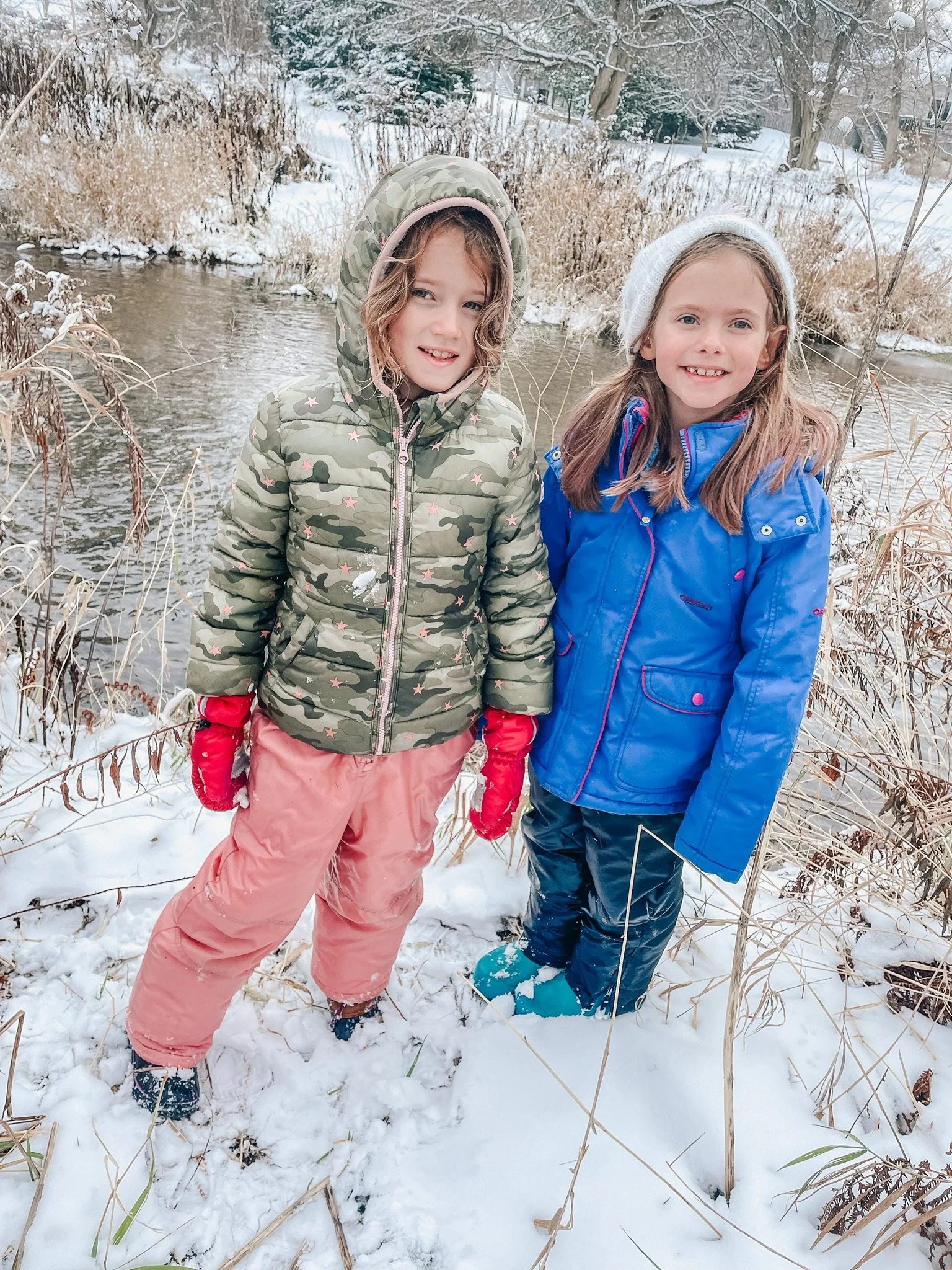 Two young girls standing outdoors in winter snow, wearing colorful winter jackets and gloves, near a small river or stream with snow-covered trees and grass in the background.