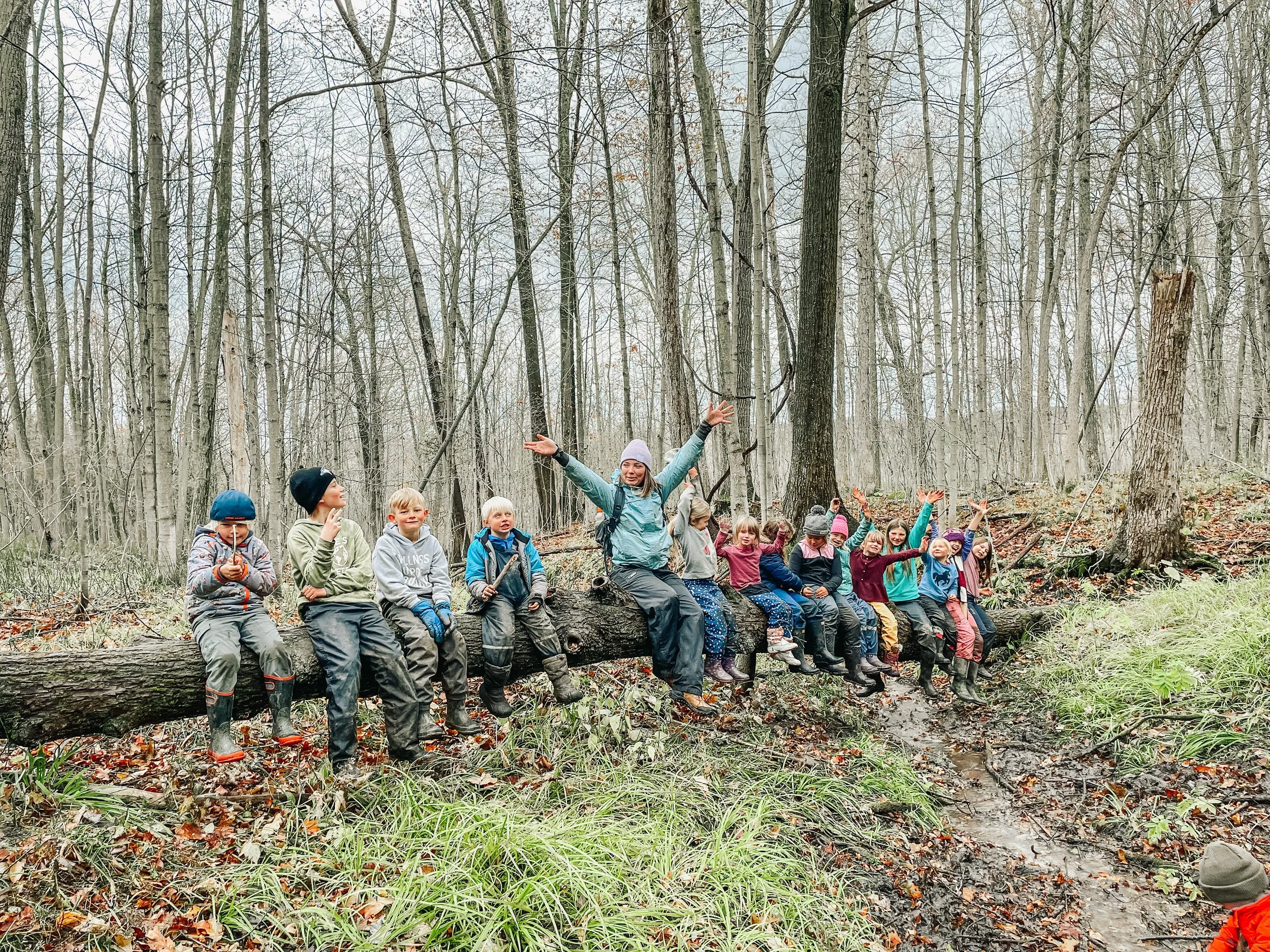 A group of children and a woman sitting on a fallen tree in a forest, some children with arms raised and smiling.
