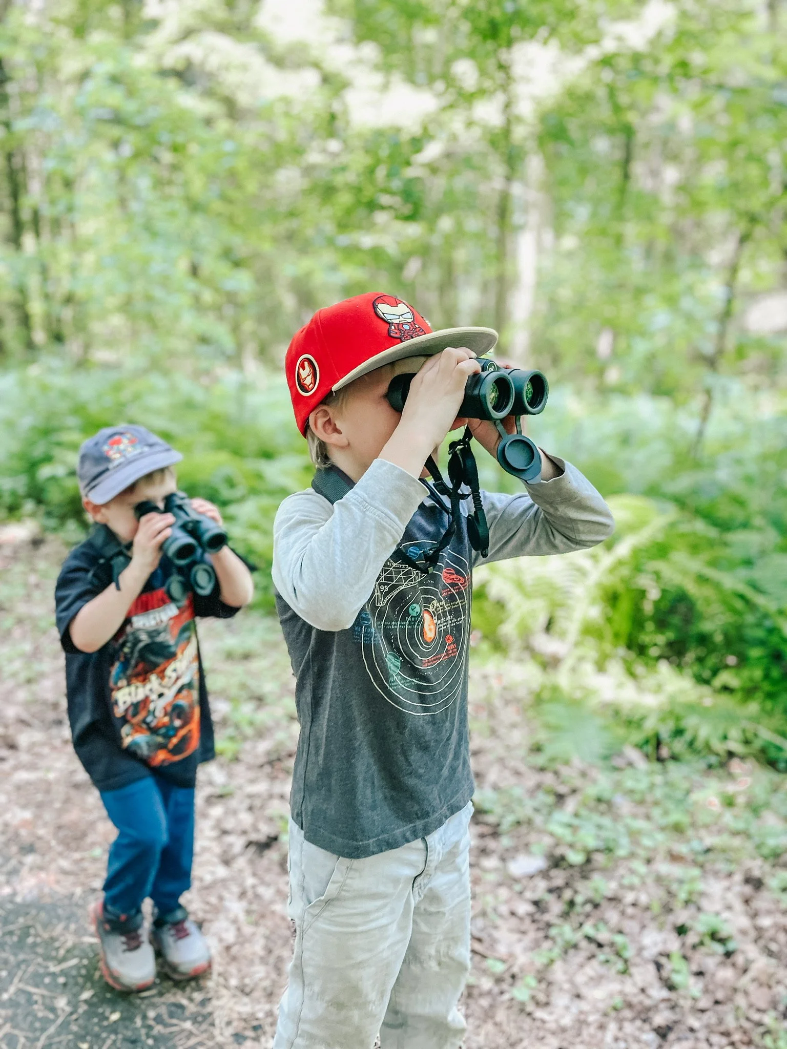 Two young boys in a forest, each looking through binoculars. The boy in the foreground wears a red cap and a gray long-sleeve shirt with beige pants, while the boy in the background wears a gray cap and black shirt with blue pants.