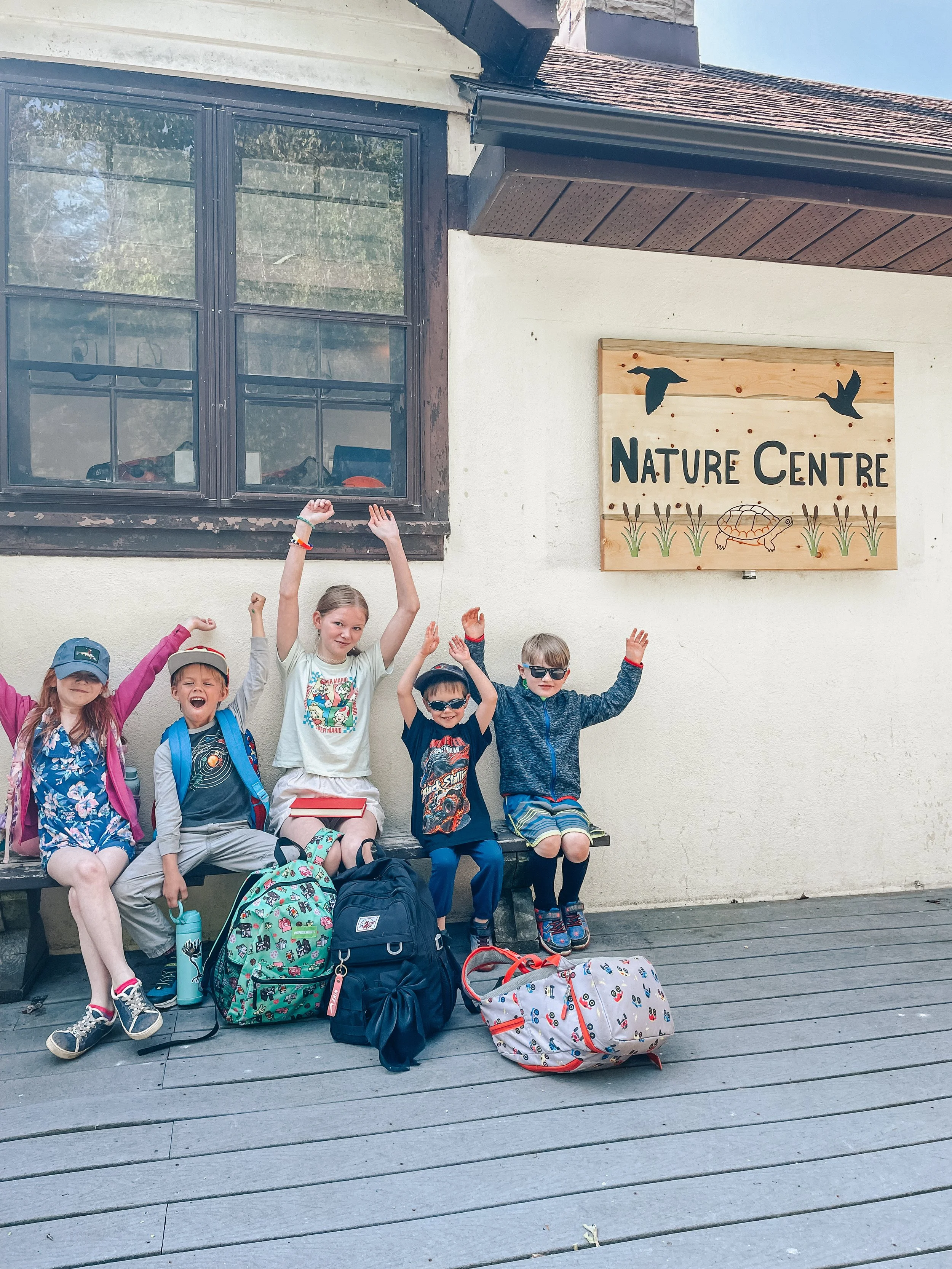 Five children sitting on a bench outside a building with a sign that reads 'Nature Centre.' They are smiling and raising their hands, with backpacks and a duffel bag in front of them.