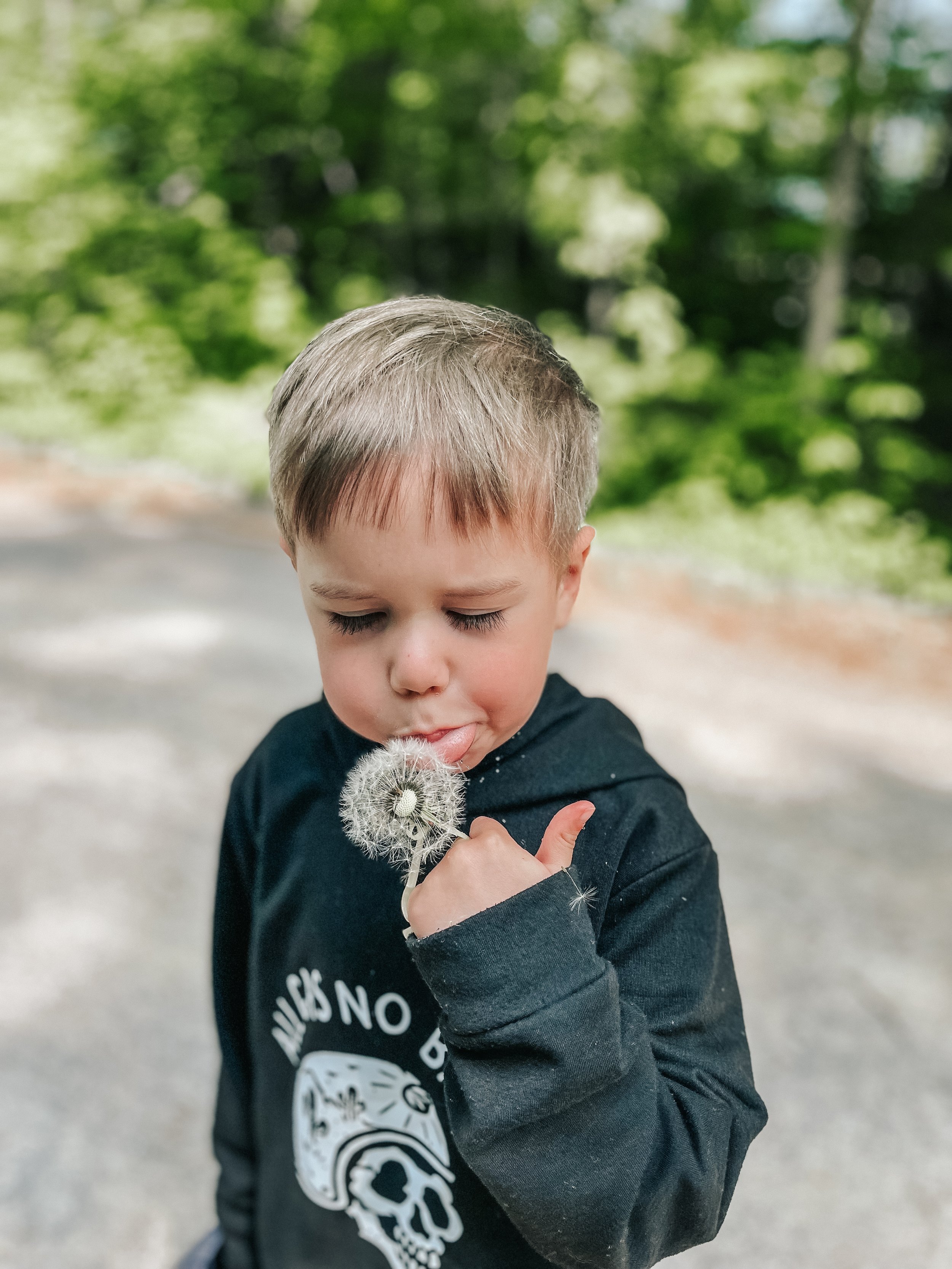 A young boy with short blond hair holding a dandelion seed head close to his face, outdoors with green trees in the background.