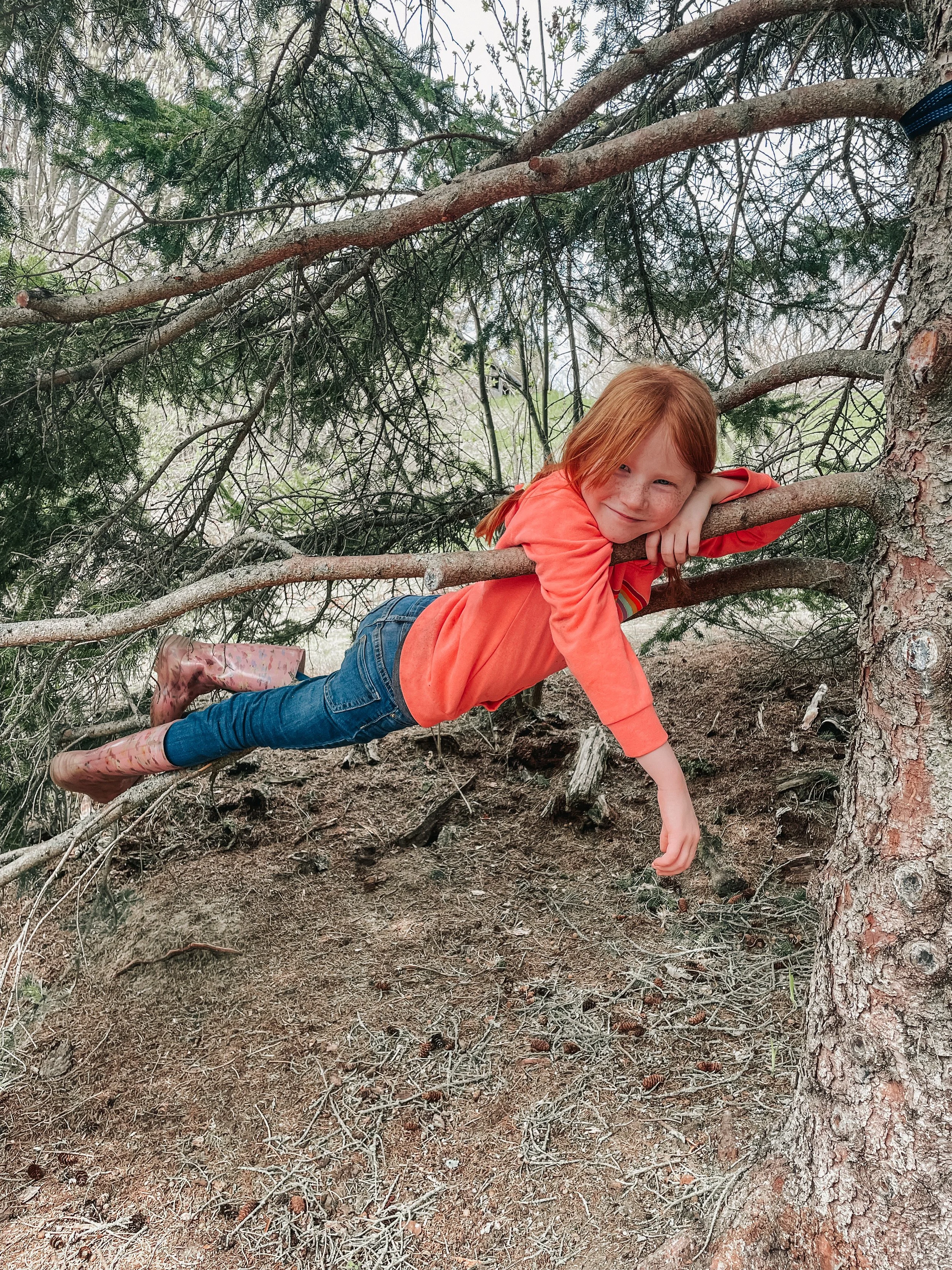 A young girl with red hair, wearing a coral long-sleeve shirt and rain boots, is lying on a tree branch in an outdoor wooded area, smiling at the camera.