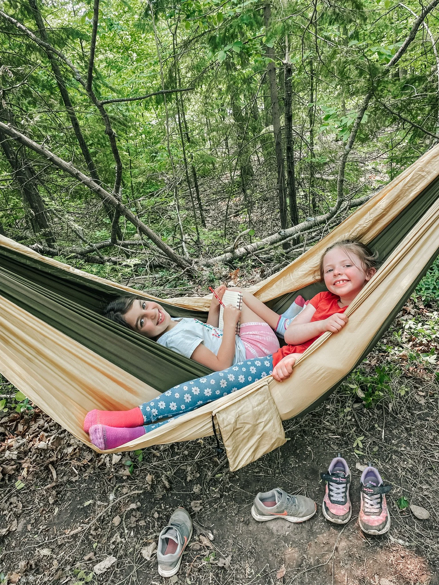 Two young girls lying in a hammock in a green forest, smiling at the camera. Shoes are placed on the ground nearby.