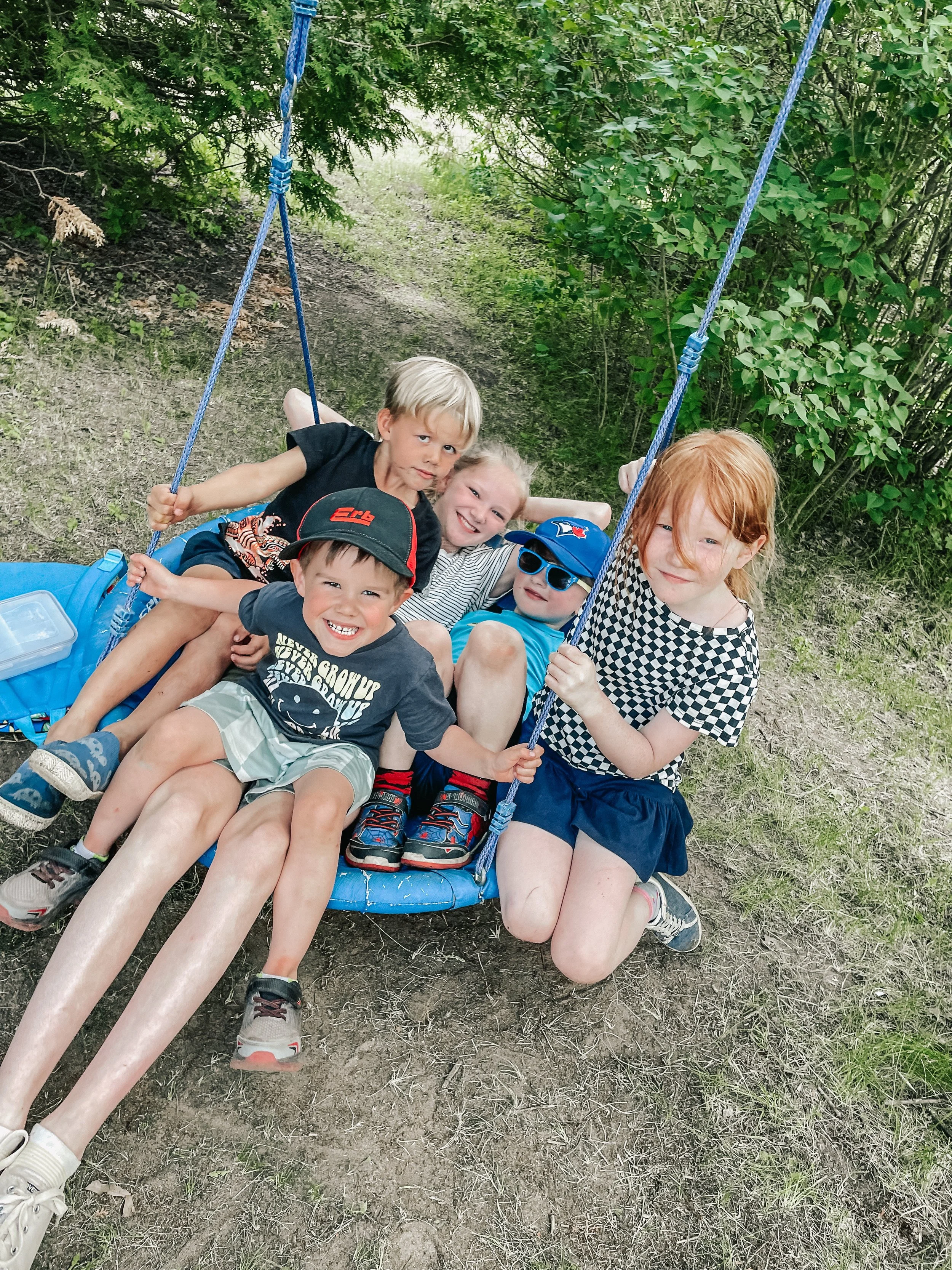 Group of five children playing on a blue swing in an outdoor park area with grass and bushes.