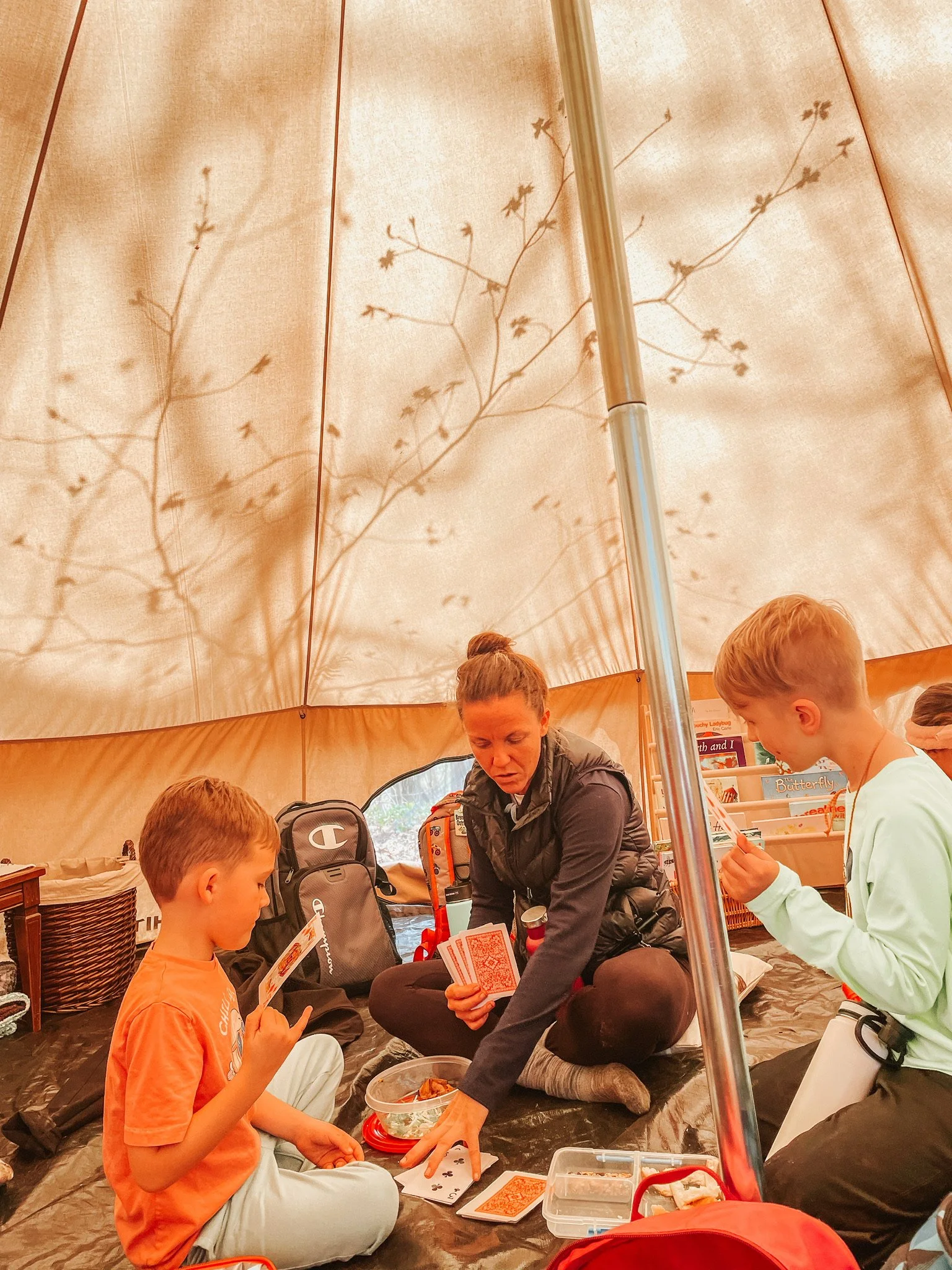 A group of children and an adult playing a card game inside a beige-colored tent.