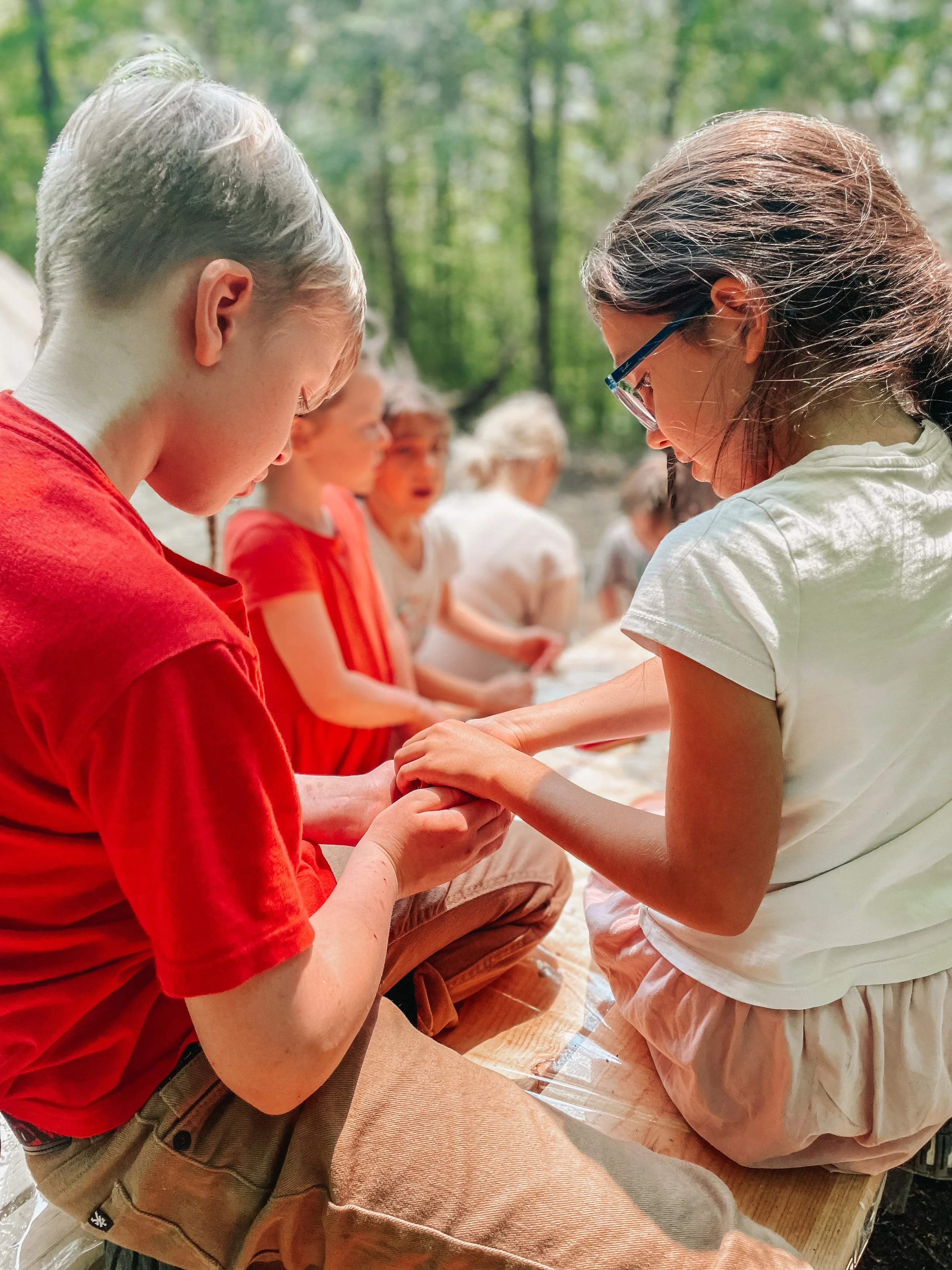 Children seated outdoors on a wooden platform, engaged in a hands-on activity, with green trees in the background.