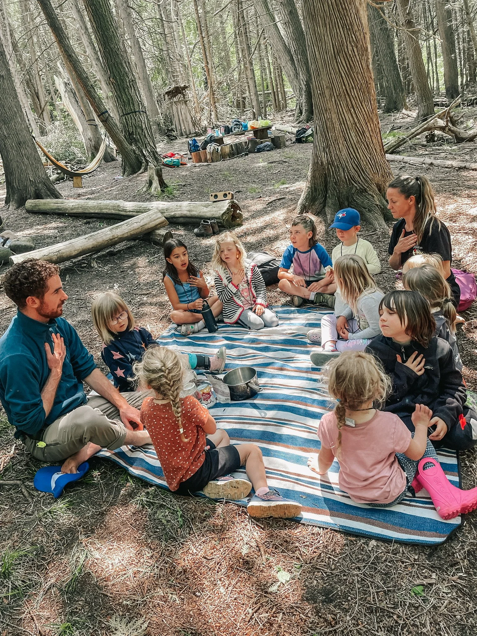 A group of children and two adults sitting on a striped blanket in a forest, engaging in a group activity.