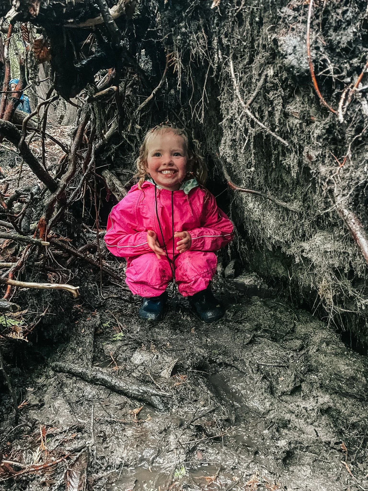 A young girl wearing a pink raincoat and black boots, smiling and squatting in a muddy, root-filled hollow in a forest.